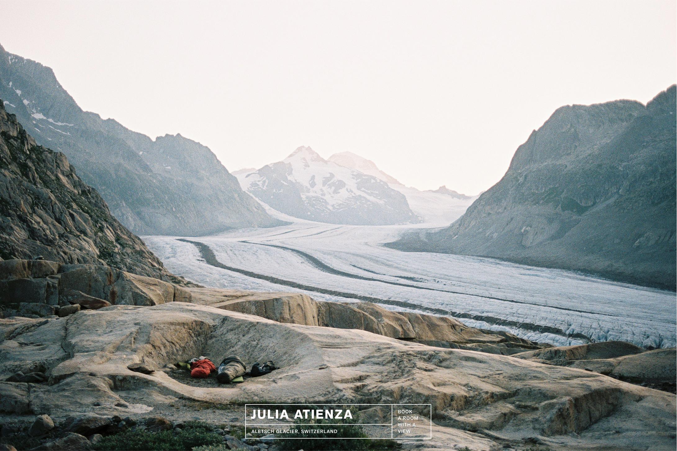 A glacier winds between mountains with Mammut sleeping bags and mountaineering gear on rocky ground in the foreground.