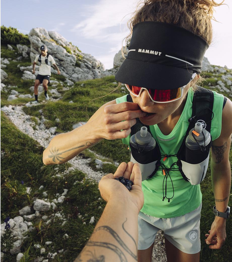 A woman trail runner, wearing Mammut gear, eats berries from a hand while another runner approaches on a rocky alpine mountain path.