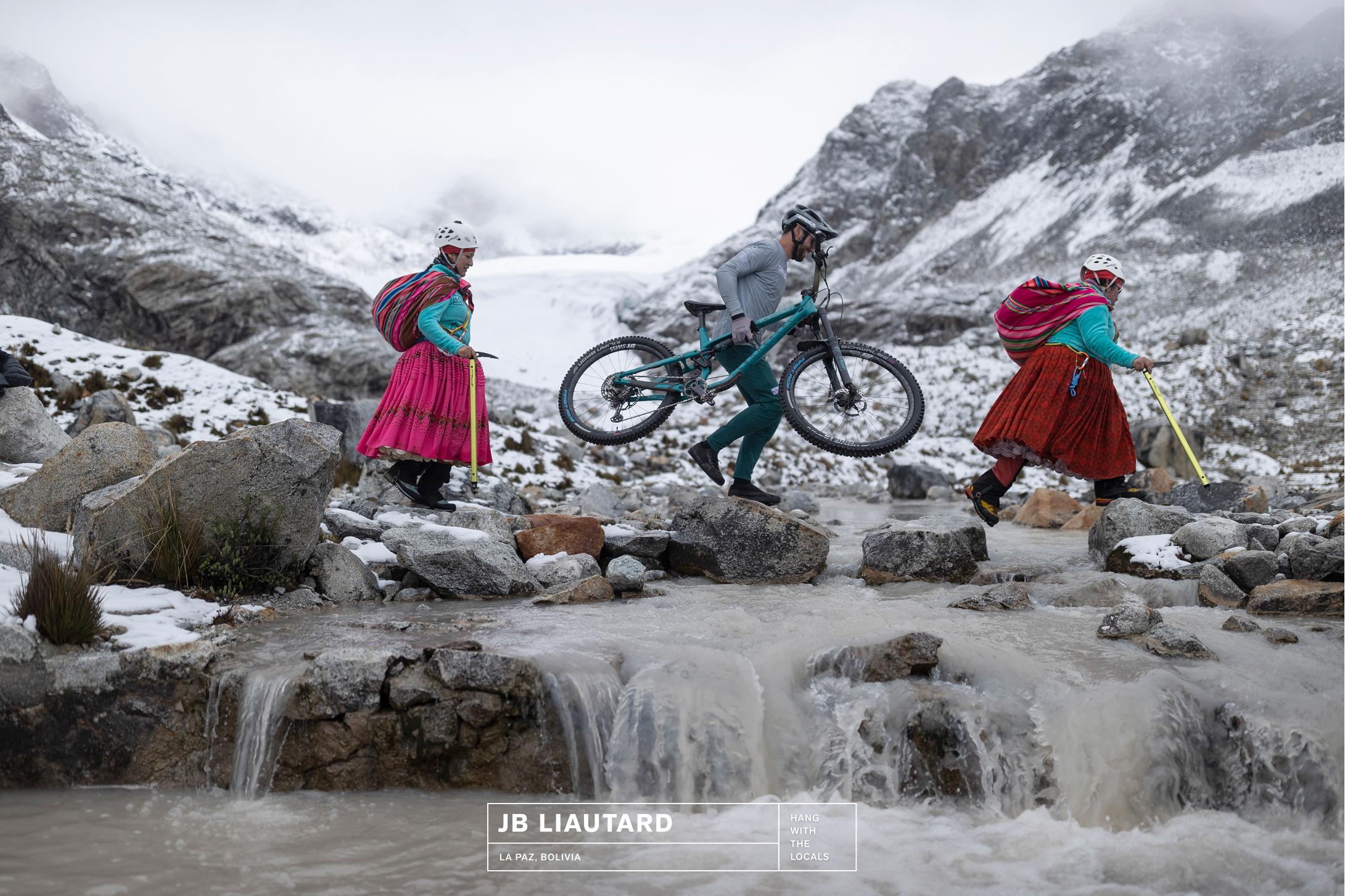Three people in traditional gear cross a rocky stream with bikes in snowy alpine mountains, showcasing Mammut adventure spirit.