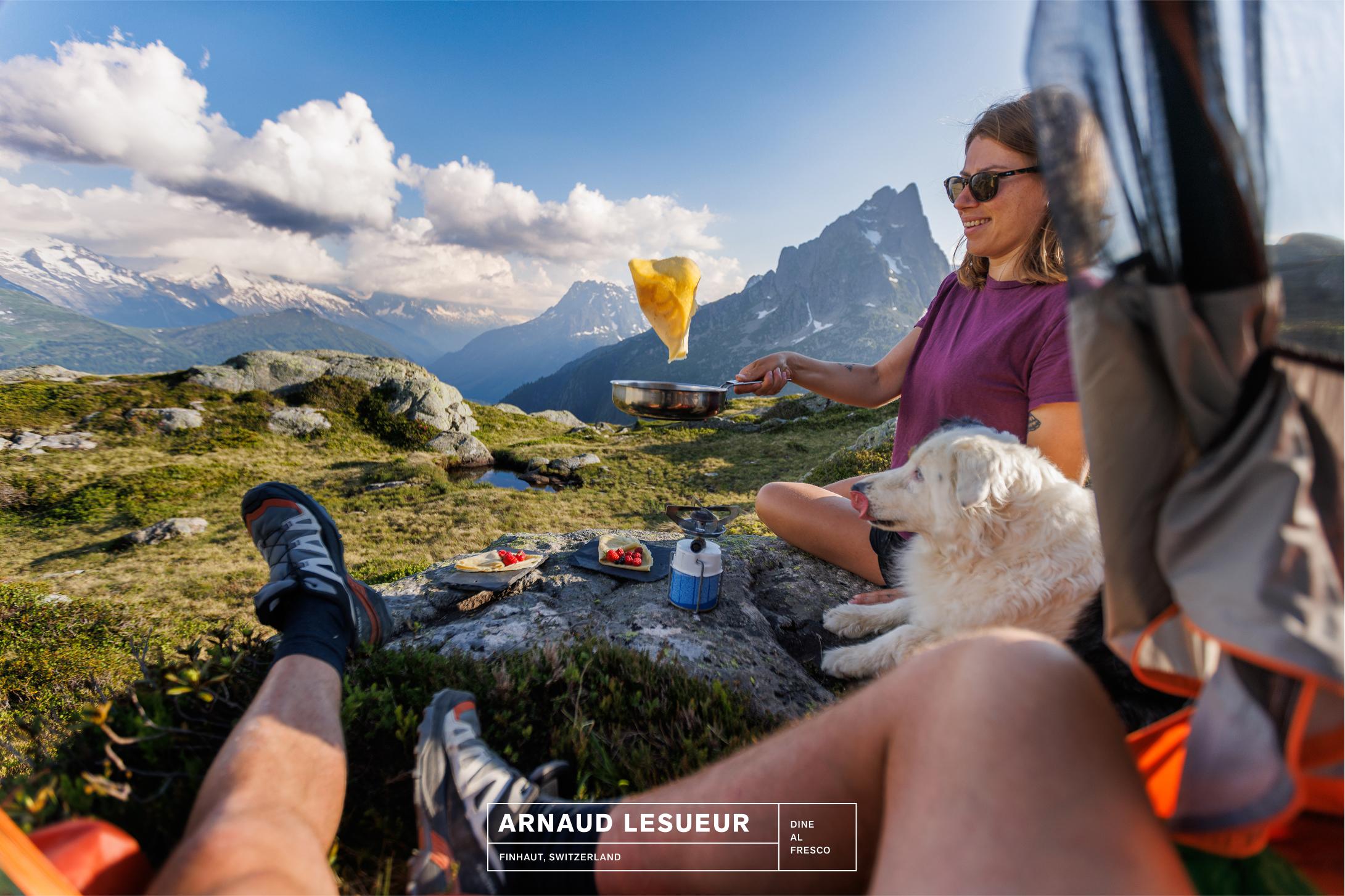 A woman in Mammut gear flips a pancake by a mountain tent, with her dog and another hiker relaxing in the alpine landscape.