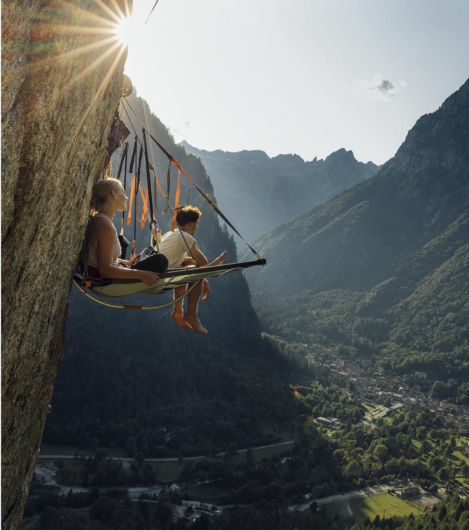 Two people relax in a Mammut hammock tent suspended on a cliff, overlooking a scenic mountain valley at sunset.