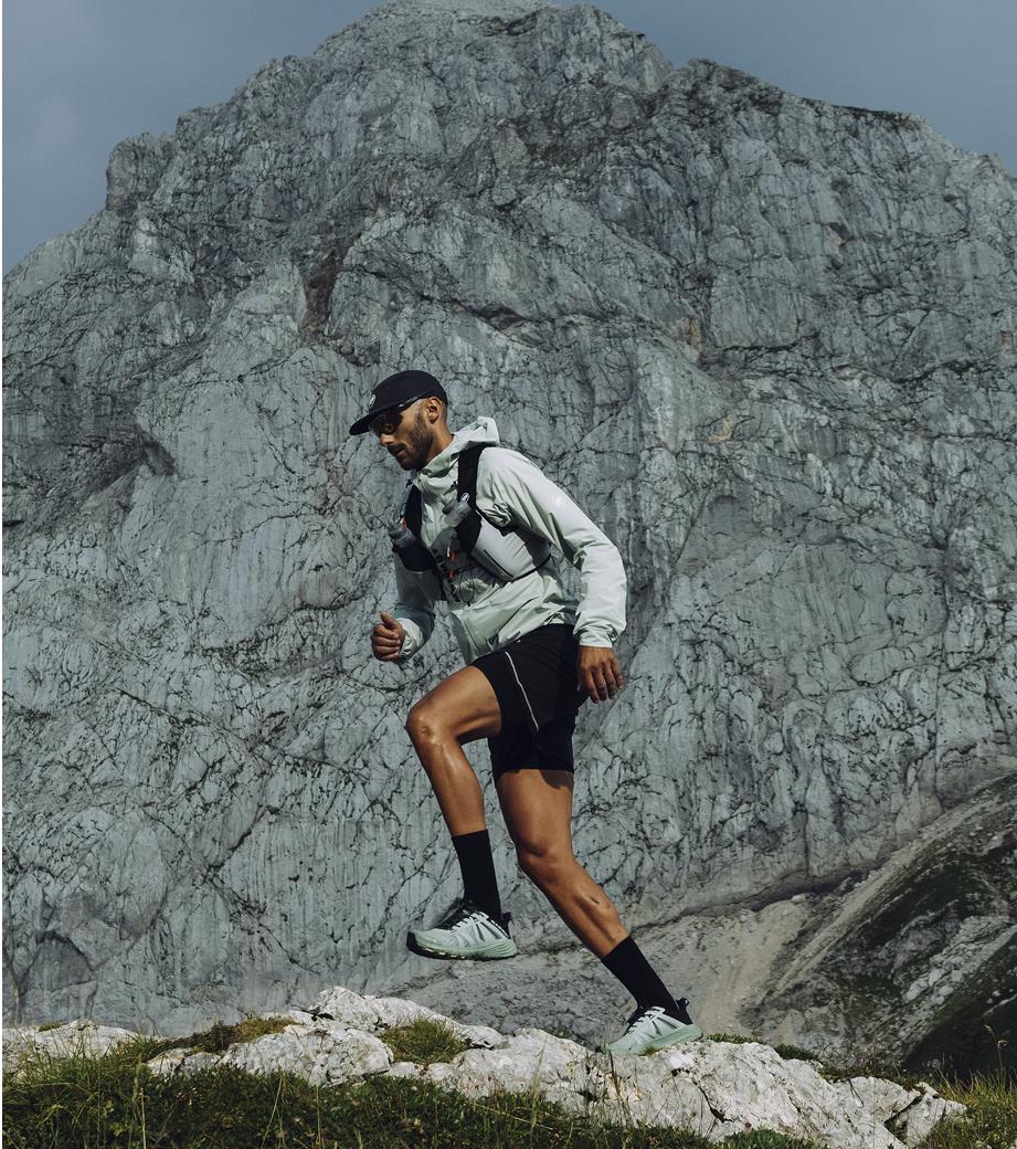 Man wearing Mammut hiking gear trekking uphill on rocky terrain with a tall, rugged mountain in the background under cloudy skies.