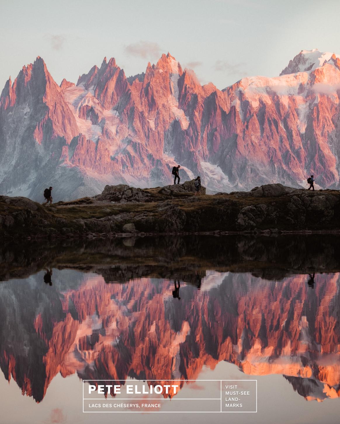 Three hikers in Mammut gear trek rocky terrain, mirrored in a calm alpine lake beneath pink-lit mountain peaks at sunrise.