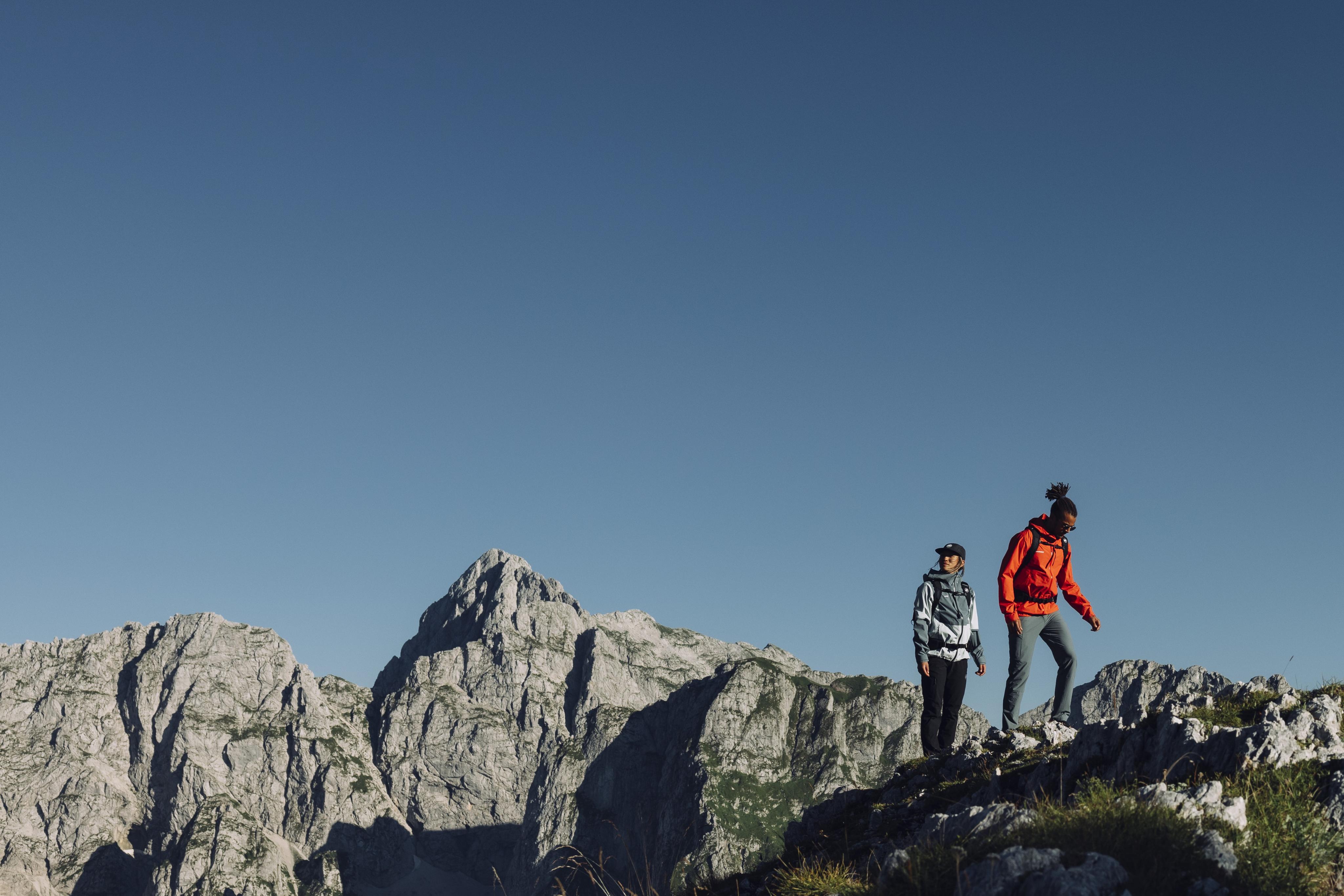 Two hikers wearing Mammut outdoor gear stand on a rocky mountain ridge, surrounded by rugged alpine peaks and a clear blue sky in the background.