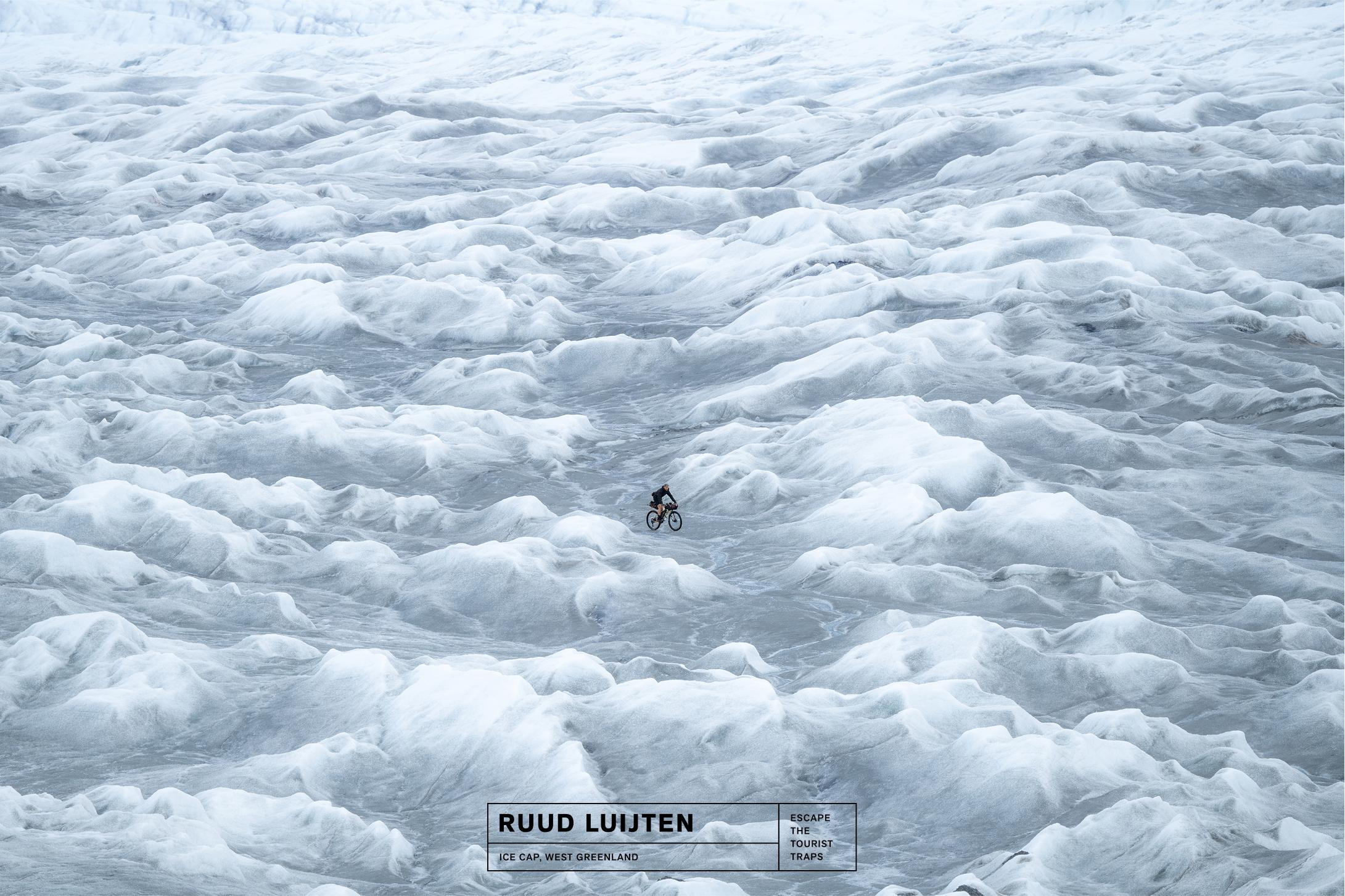 A cyclist in Mammut gear rides alone across a vast, snowy, and rugged wilderness landscape.