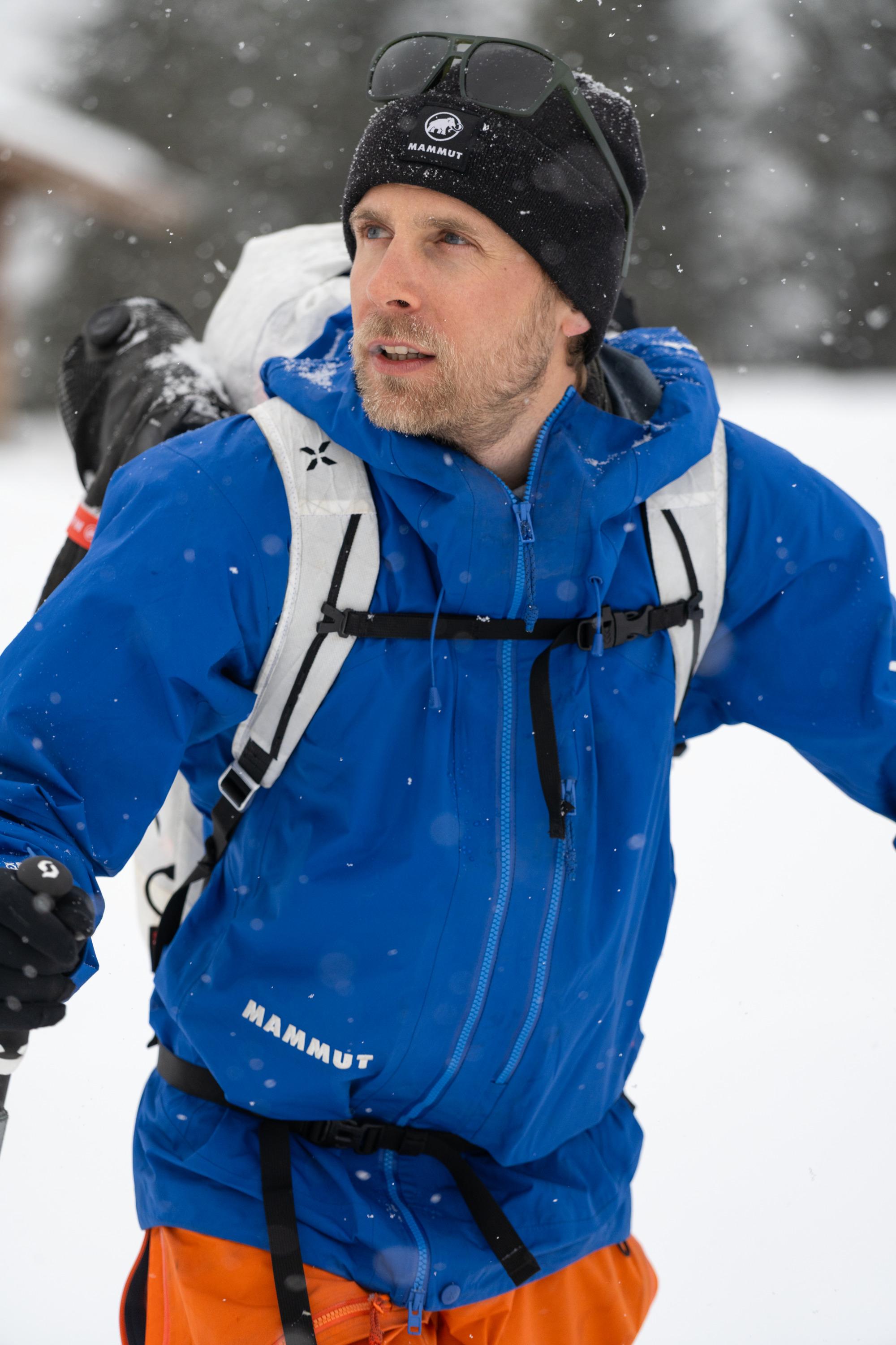 A man in a blue Mammut jacket and black beanie hikes through snowy terrain, carrying a backpack with sunglasses on his hat.