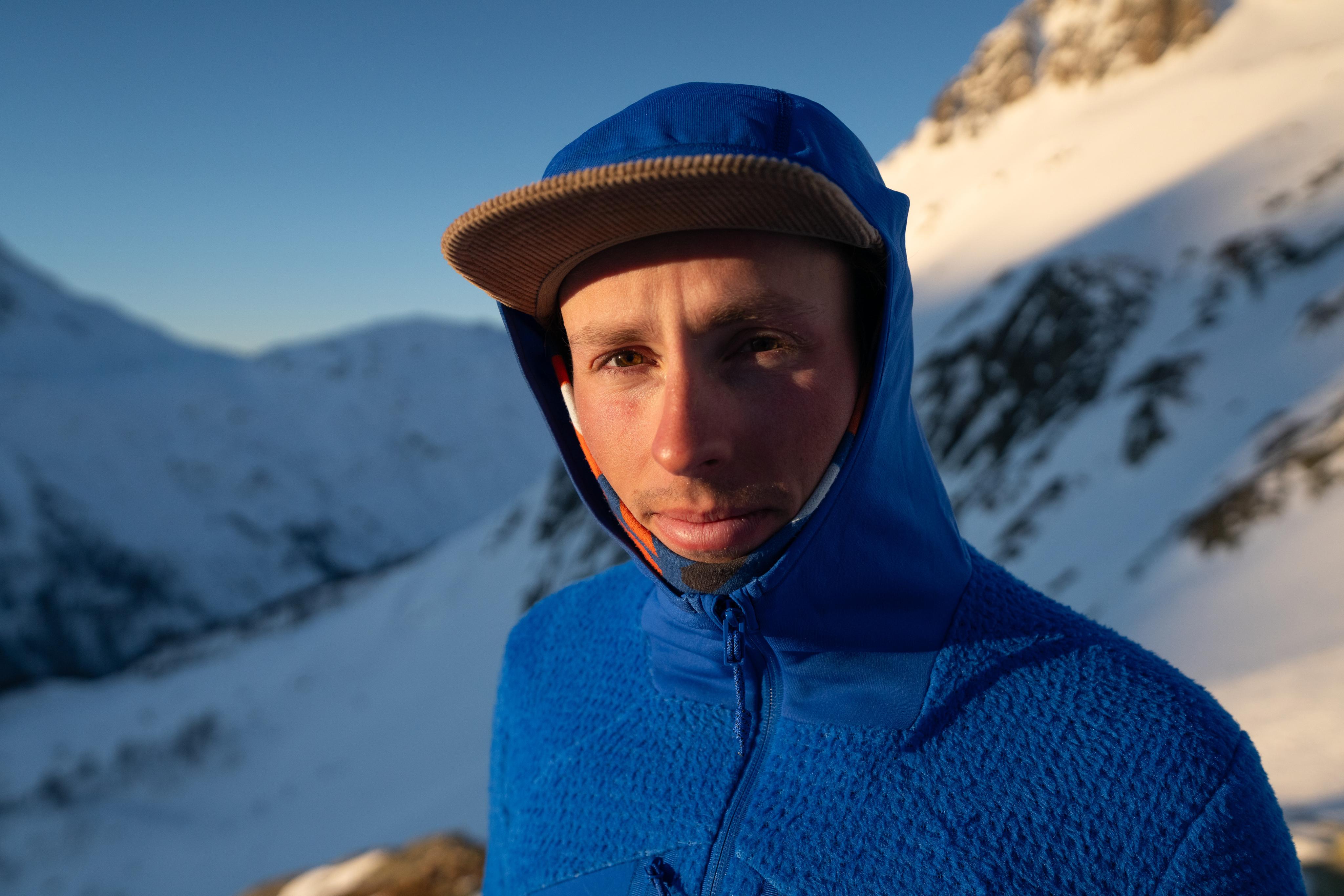 A smiling person in a Mammut blue hooded jacket and tan cap, standing outdoors in a snowy mountain landscape.
