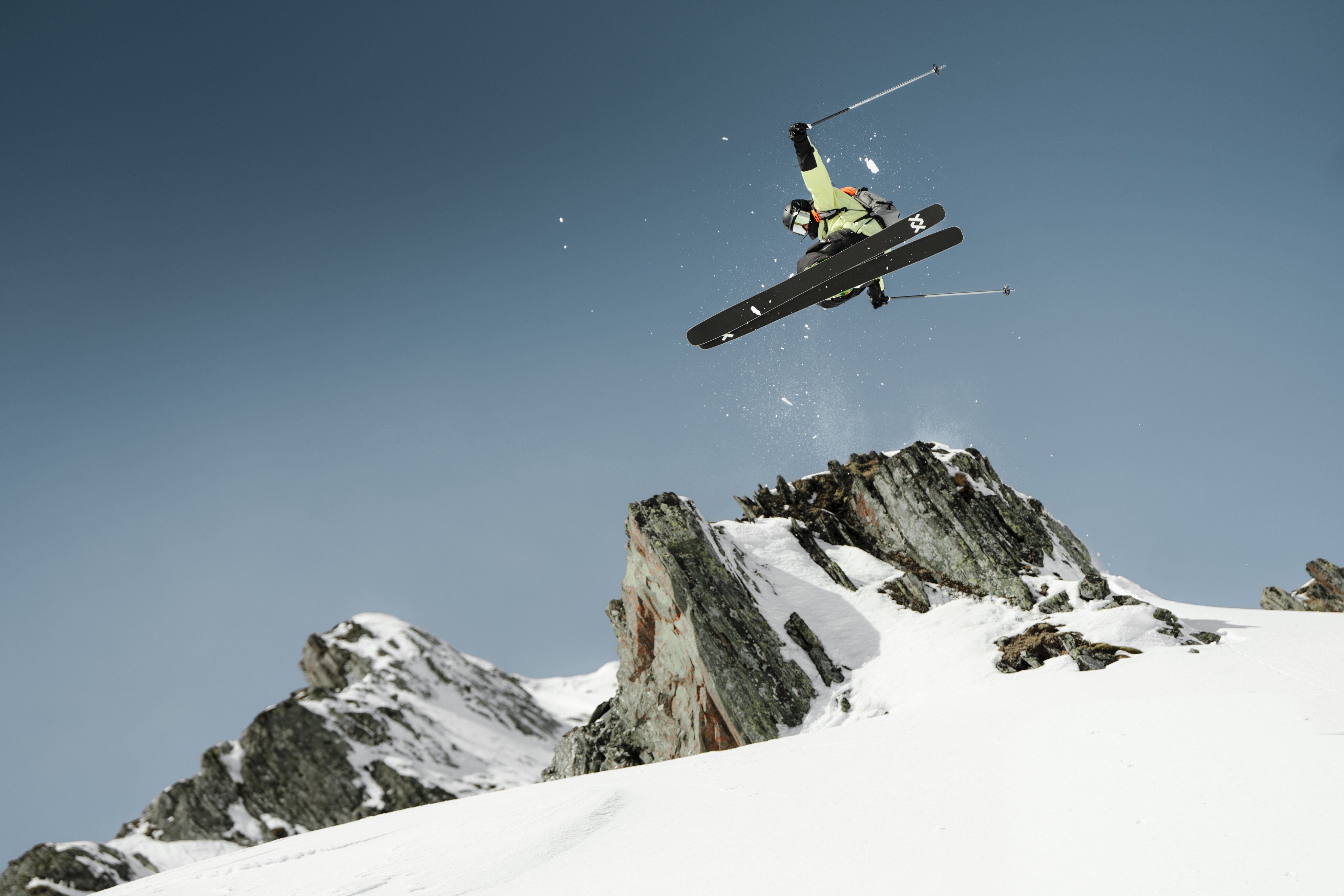 Skier in a Mammut pink jacket performing a jump off a snowy slope, set against the backdrop of a clear blue sky and majestic mountain range.
