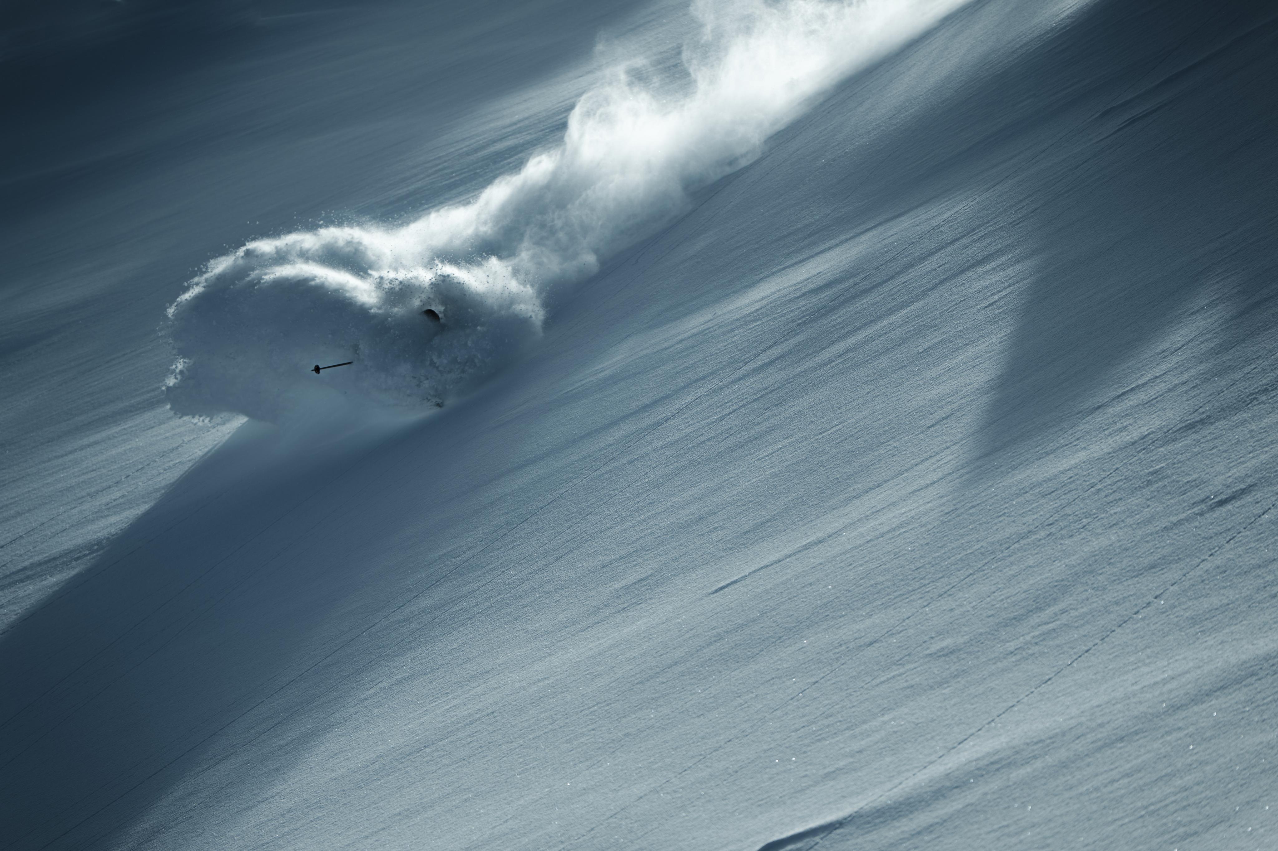A skier wearing Mammut gear expertly descends a steep, snowy slope under a clear blue sky, carving through fresh powder and leaving an impressive trail behind.