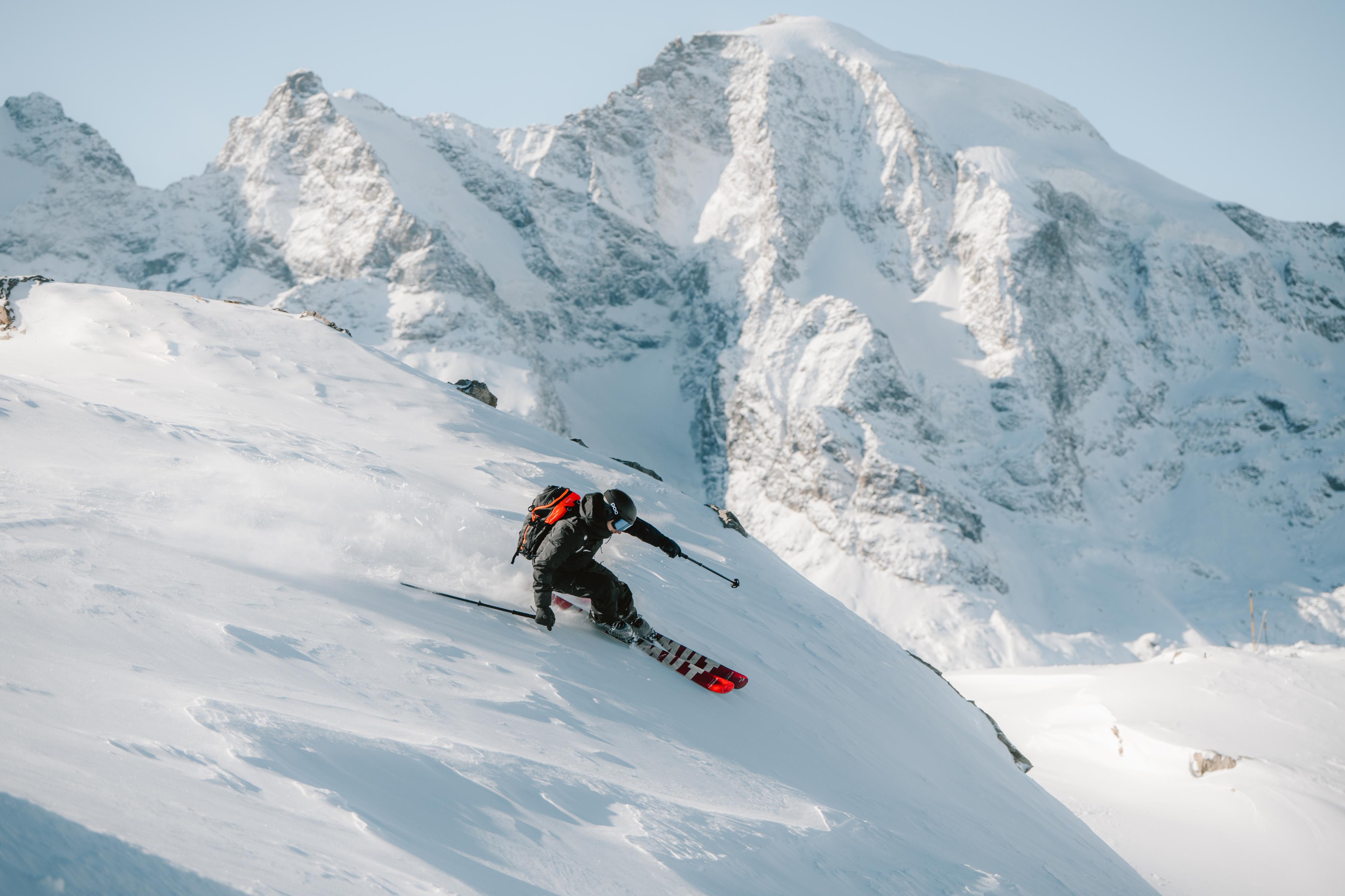 Person in pink Mammut ski gear and goggles carries skis on shoulder against a snowy mountain background.