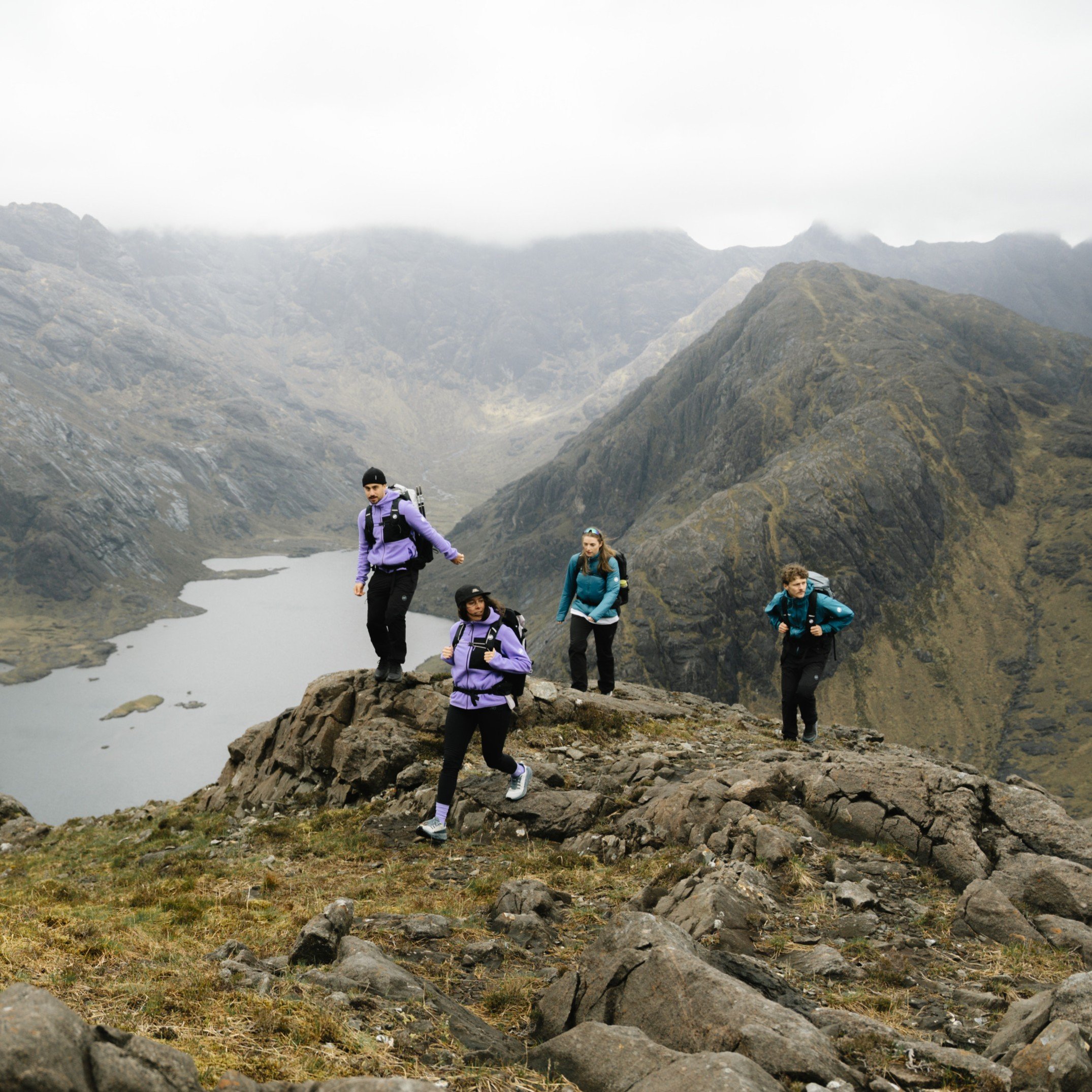 Four people in Mammut hiking gear climb a steep, rocky mountain path with misty mountains and lush greenery in the background.