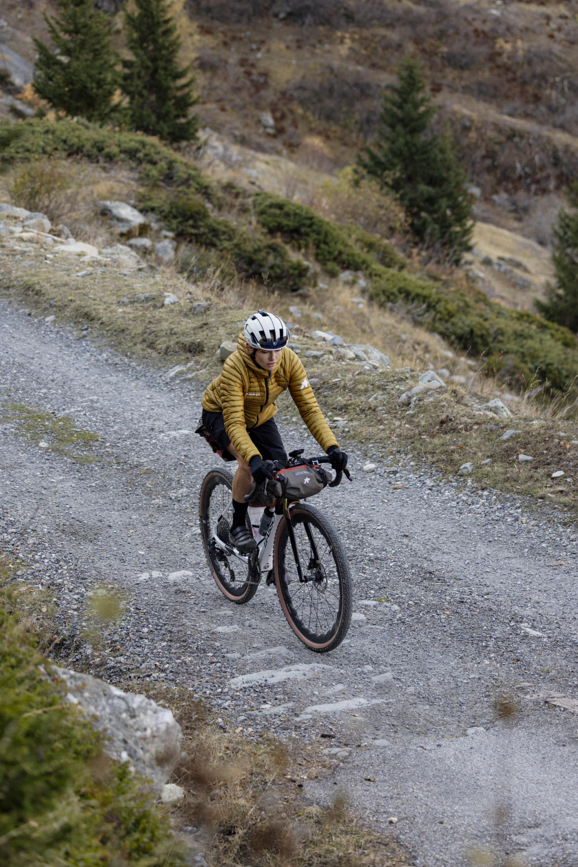 Cyclist in a Mammut yellow jacket rides a gravel road surrounded by dense trees and rugged, rocky terrain, showcasing the perfect blend of outdoor adventure and high-quality gear.