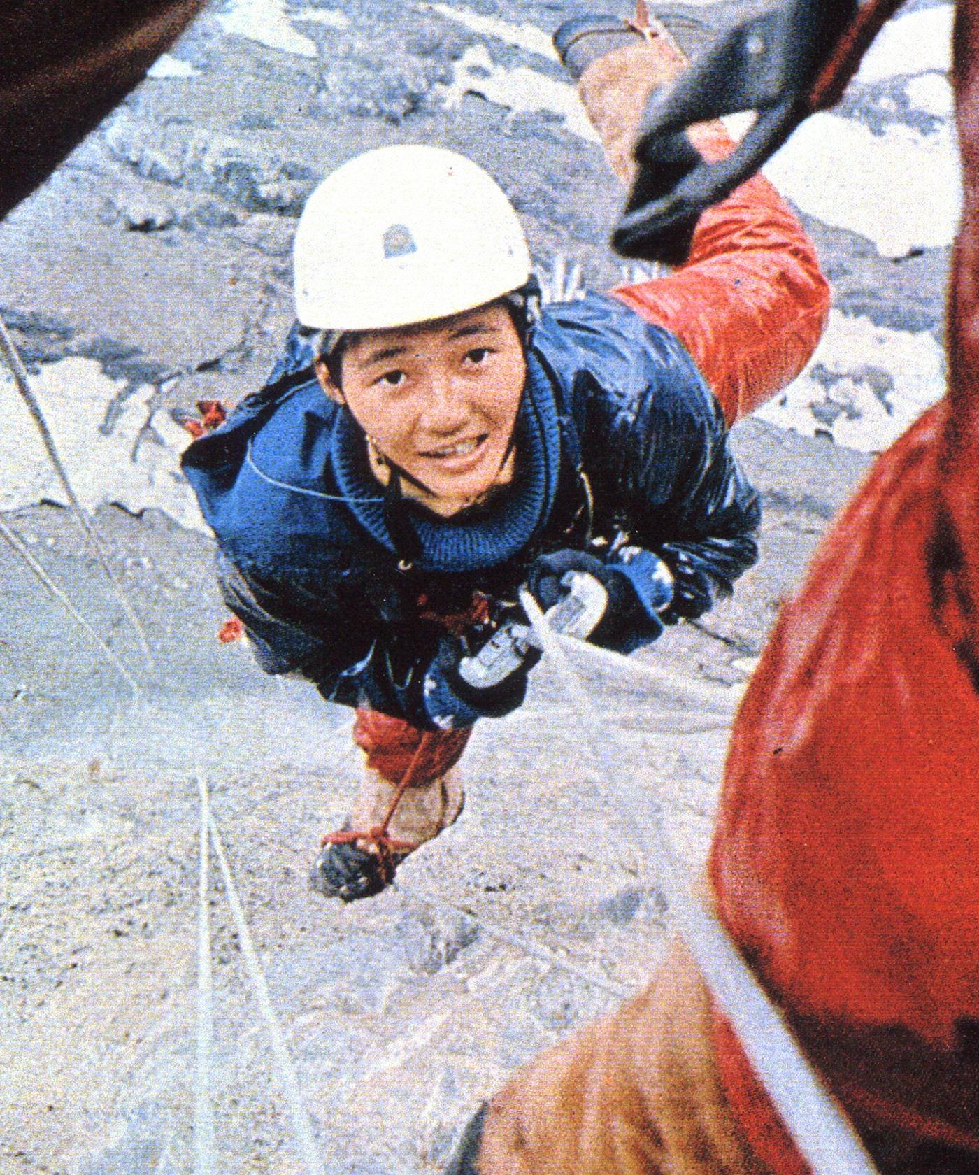 A climber wearing Mammut gear and a helmet smiles while scaling a rocky mountain, secured with ropes and protective gloves.
