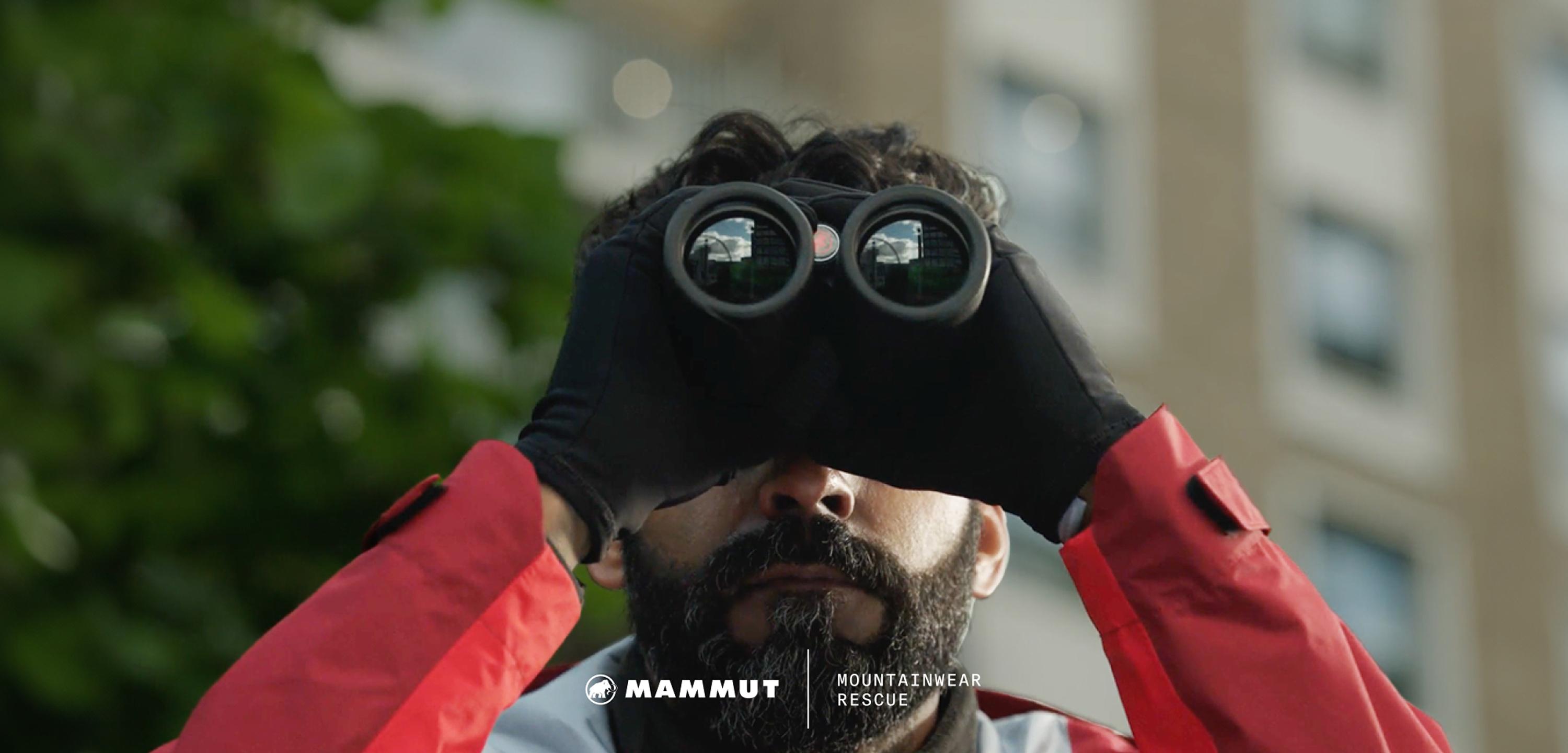 A bearded man in Mammut gloves scans the outdoors with binoculars, urban buildings softly blurred in the background.