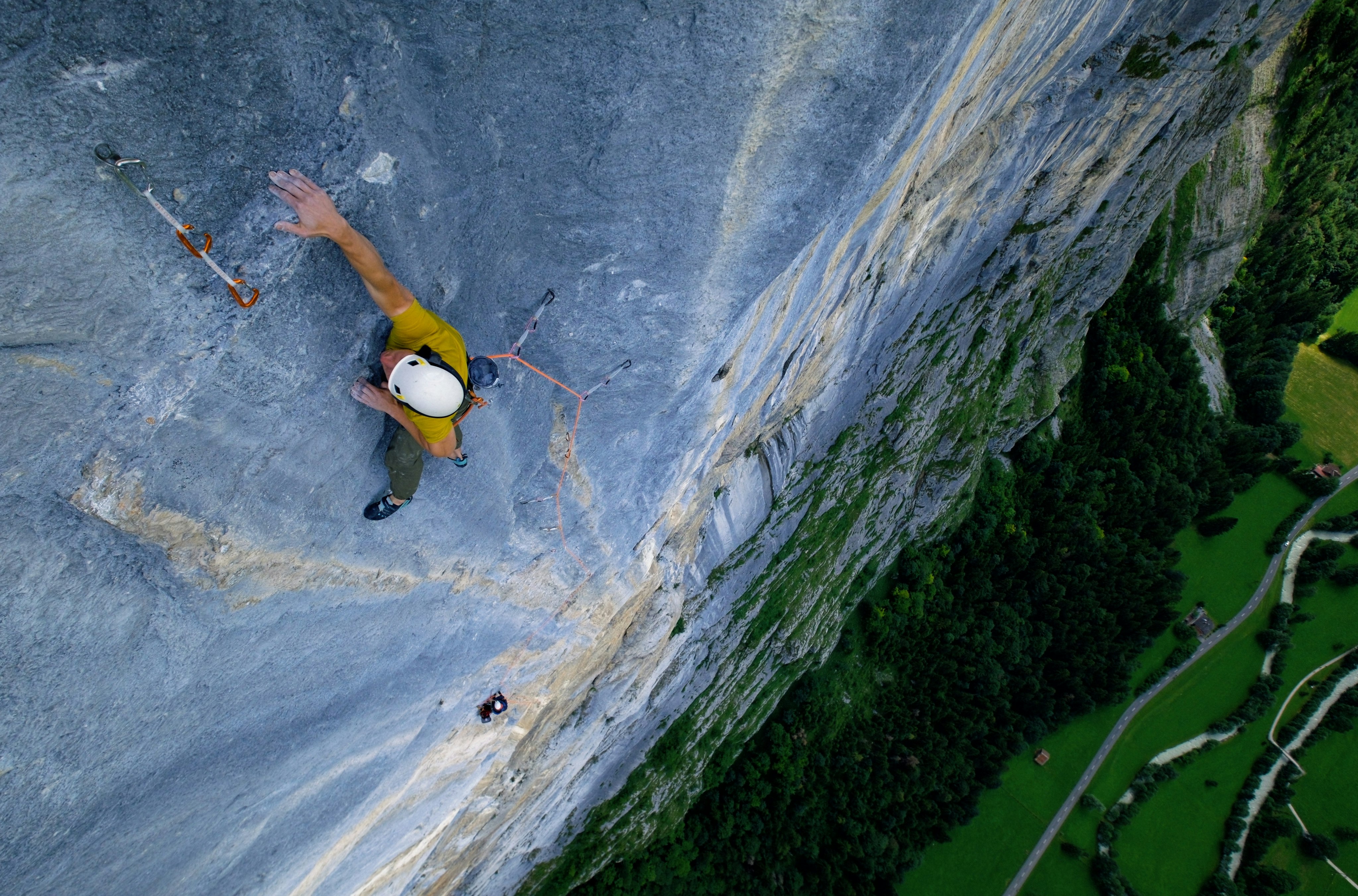 Katherine Choong, Mammut rock climber with helmet scaling a steep cliff, surrounded by lush greenery and winding roads below, viewed from above.