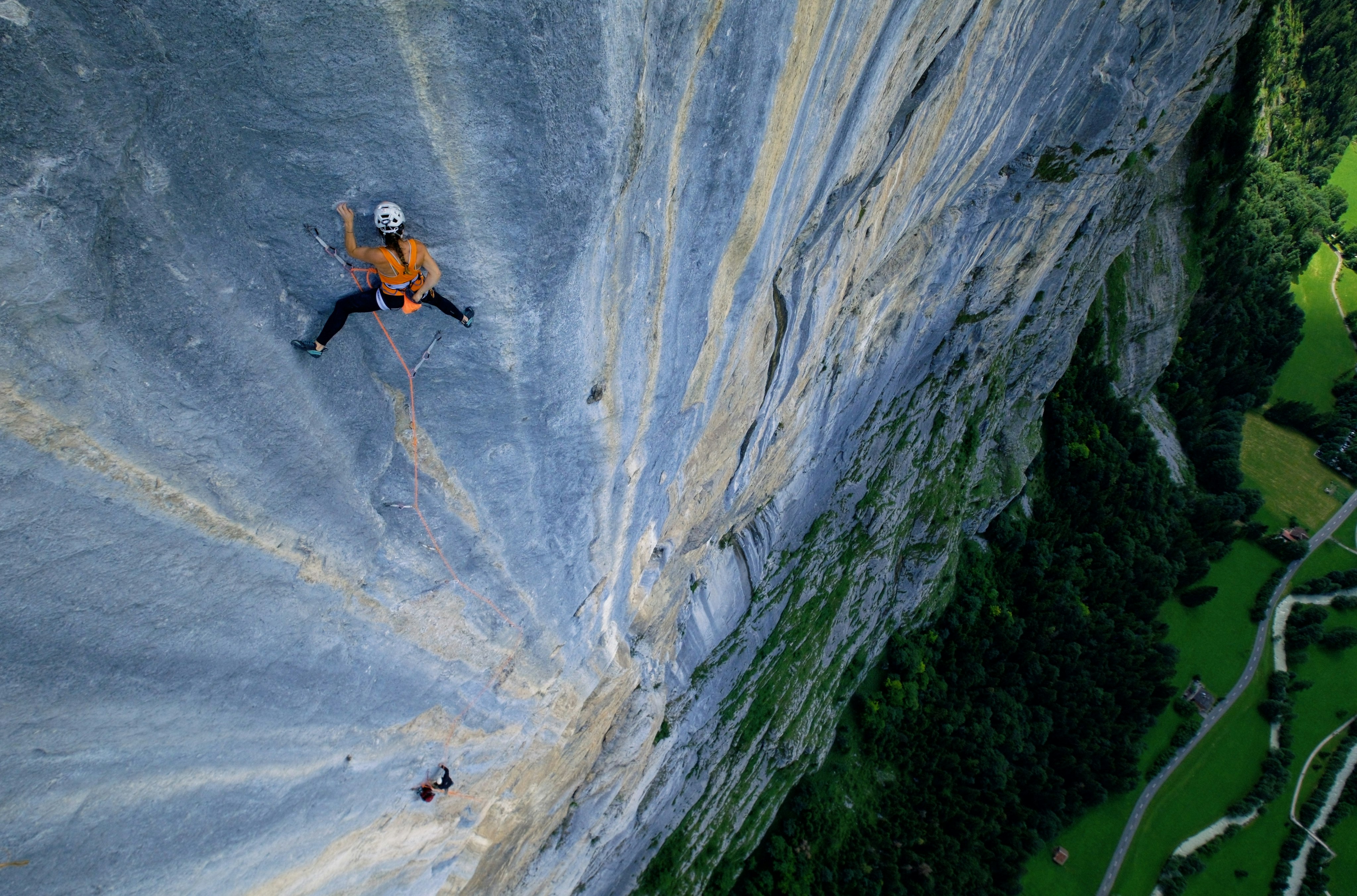 A Mammut climber in an orange shirt ascends a steep rock face, secured by high-quality Mammut ropes, with a lush forested valley stretching far below.