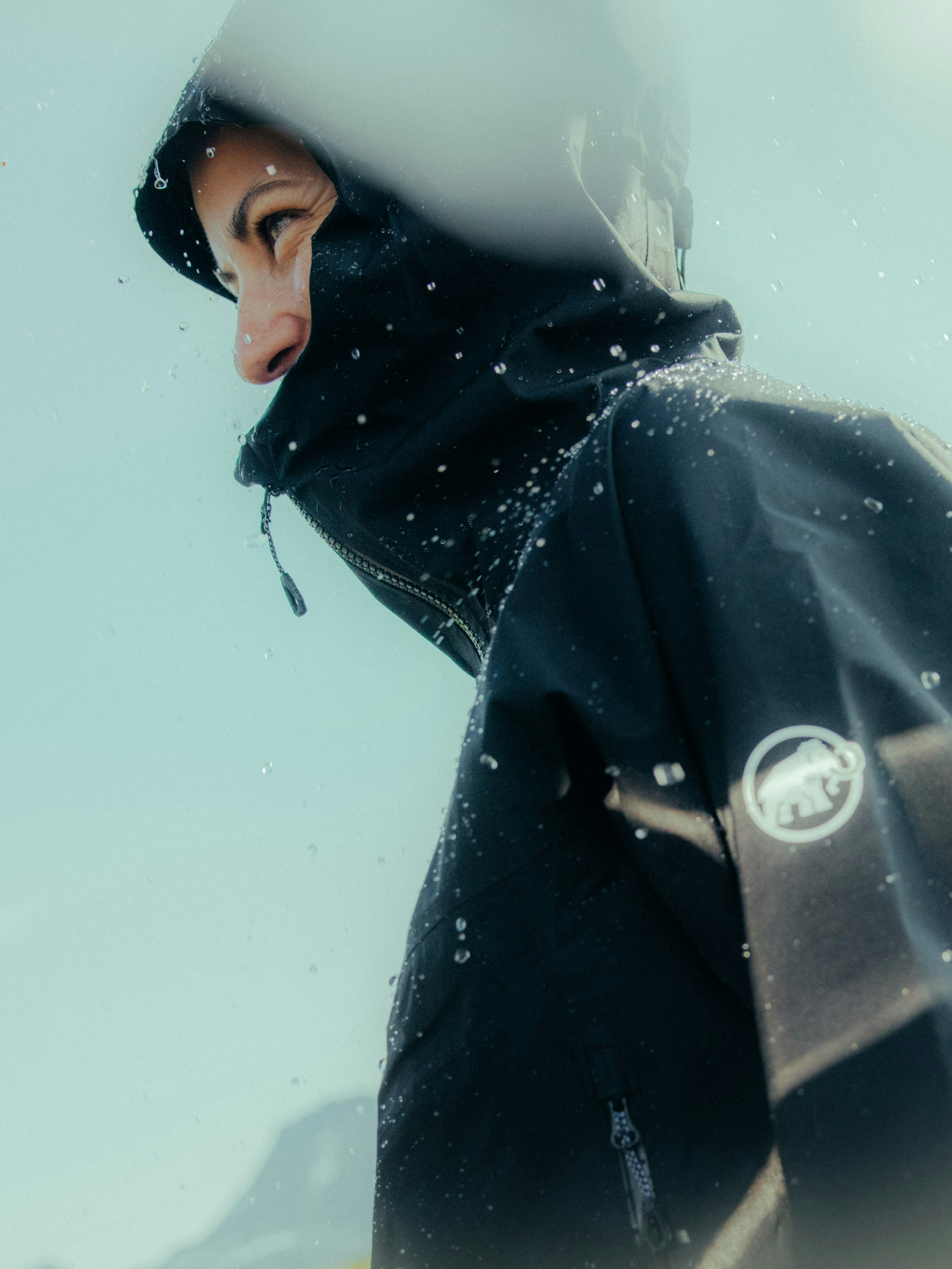 A person in a Mammut waterproof jacket with a hood is outside on a rainy day, showcasing water droplets on the high-quality fabric.