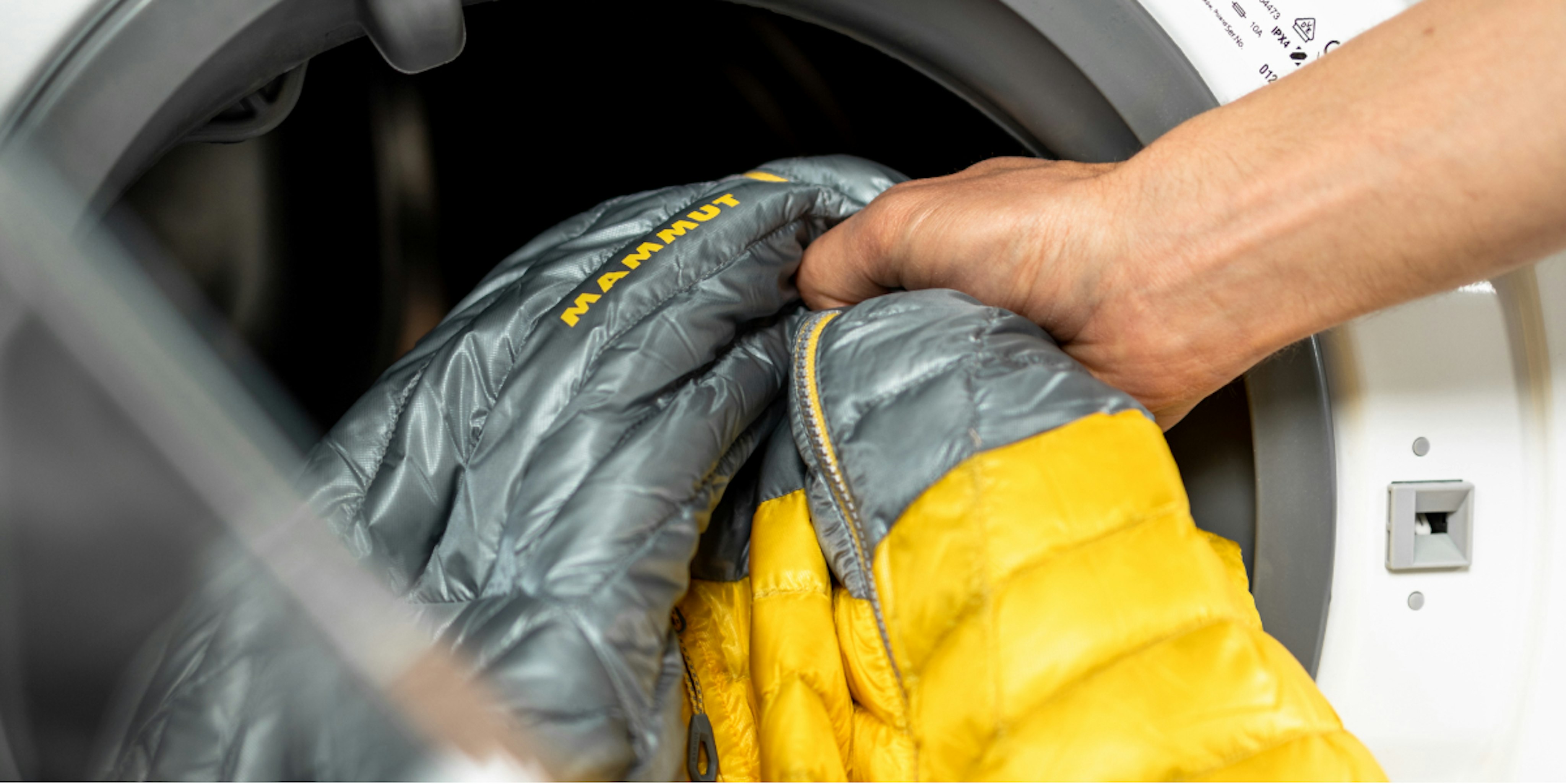 Person washing a yellow and gray Mammut jacket in a washing machine.