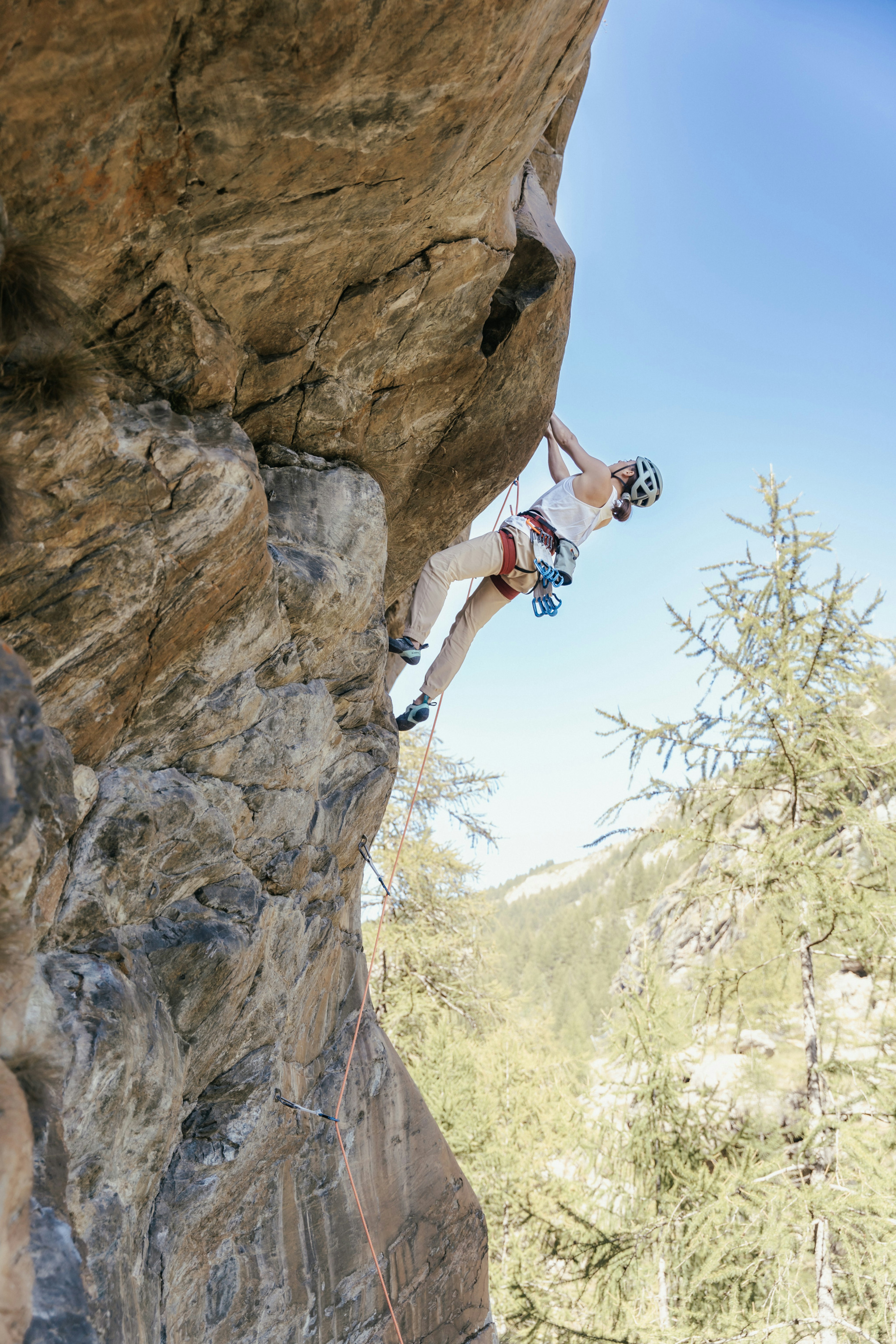 A rock climber equipped with a Mammut helmet and harness scales a steep cliff, framed by lush trees and a clear blue sky in the background.