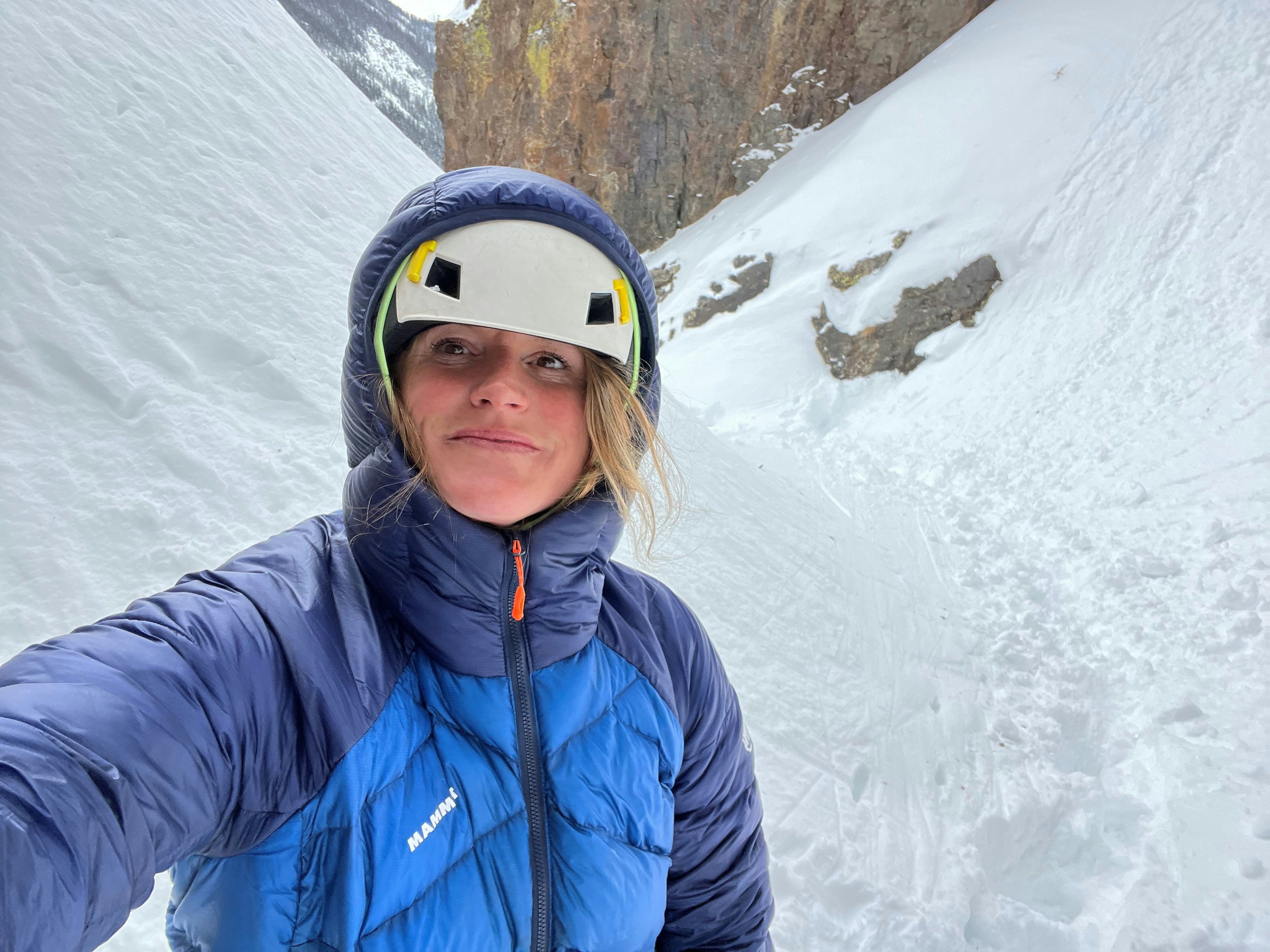 Person wearing a blue Mammut jacket and white helmet, standing in a snowy mountain pass, gazing to the side.