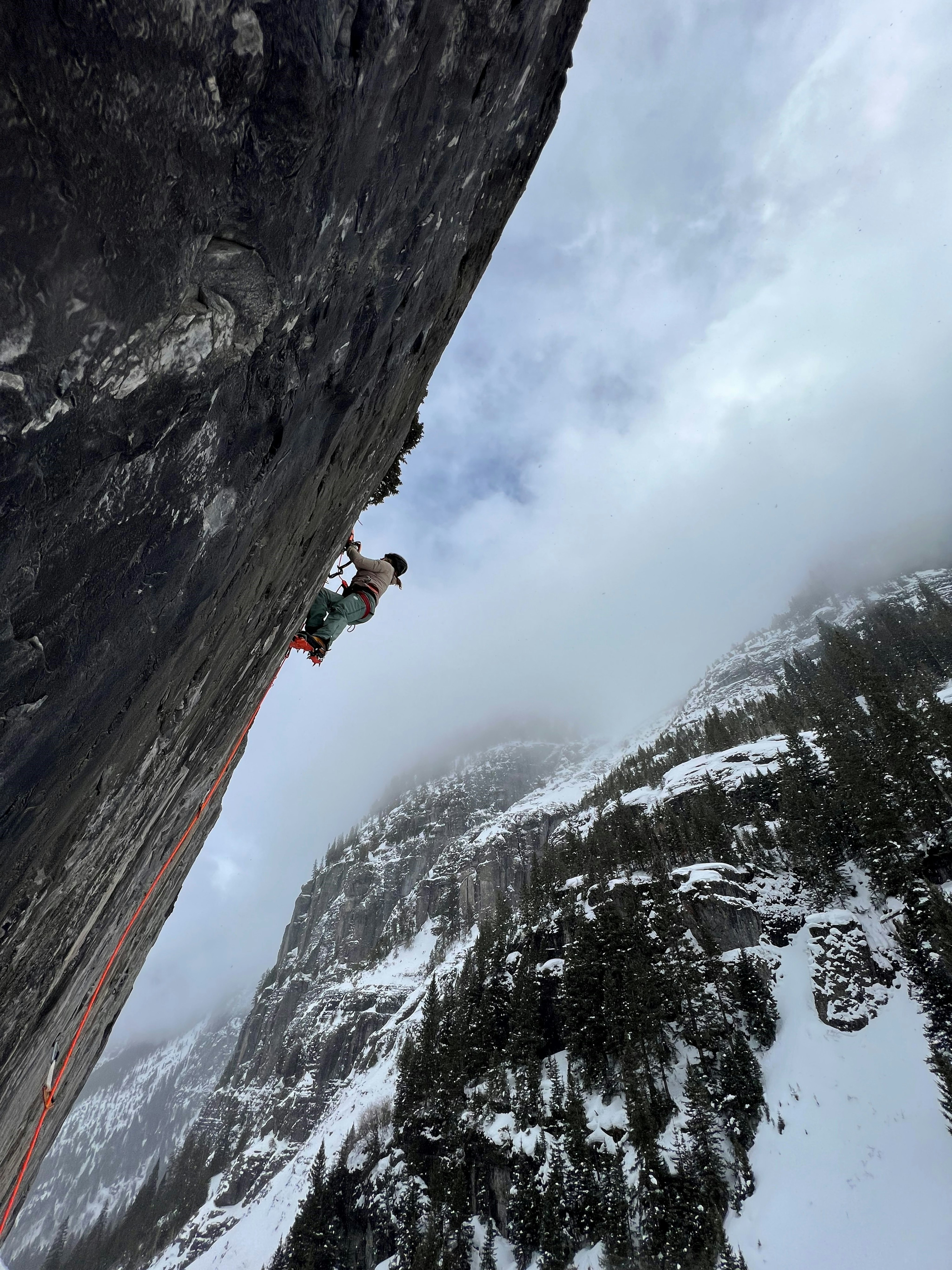 Person rock climbing on a steep cliff wearing Mammut gear, with snowy mountains and cloudy sky in the background.