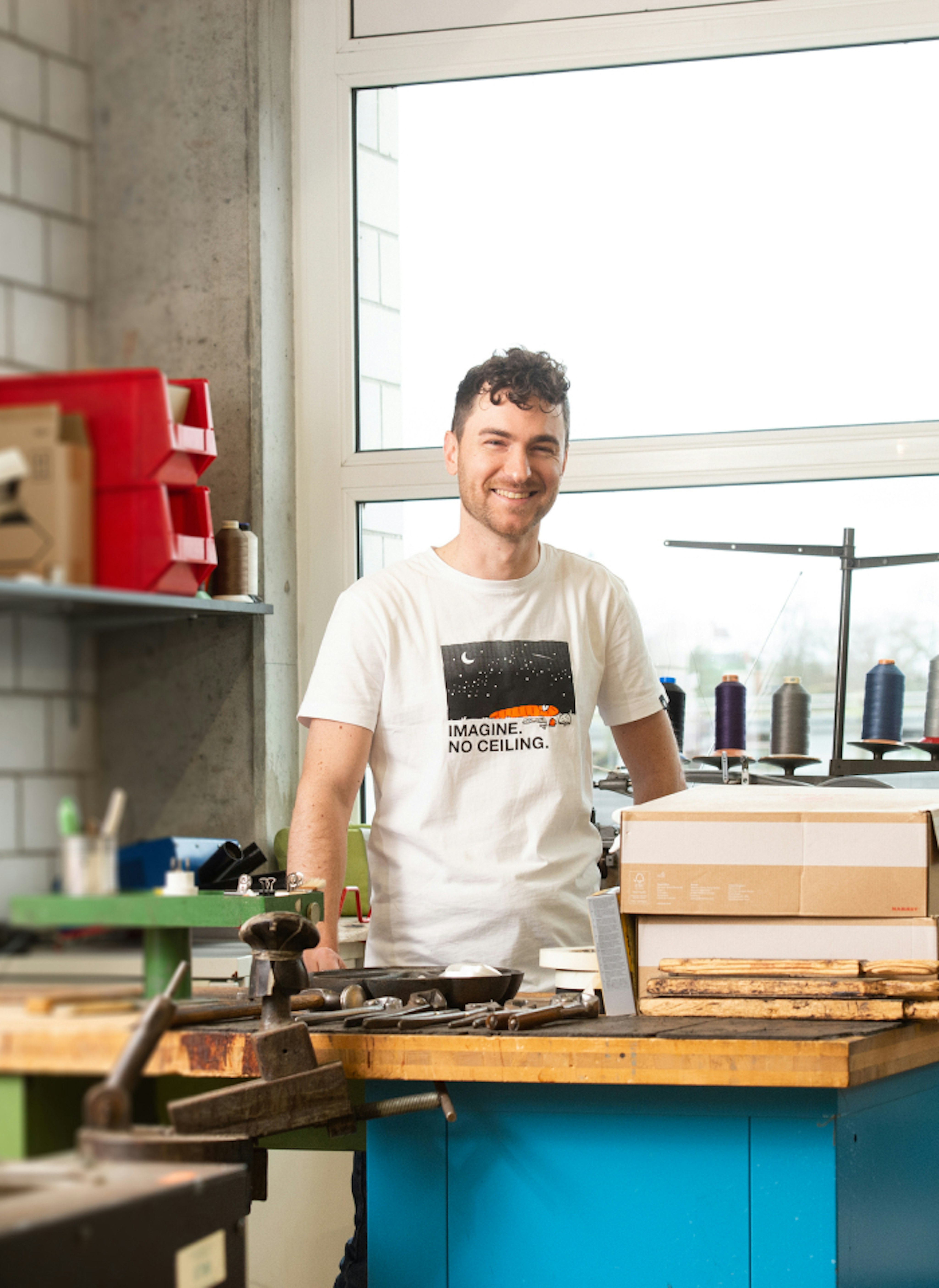 Smiling person standing in a Mammut workshop, wearing a shirt that reads "IMAGINE. NO CEILING.