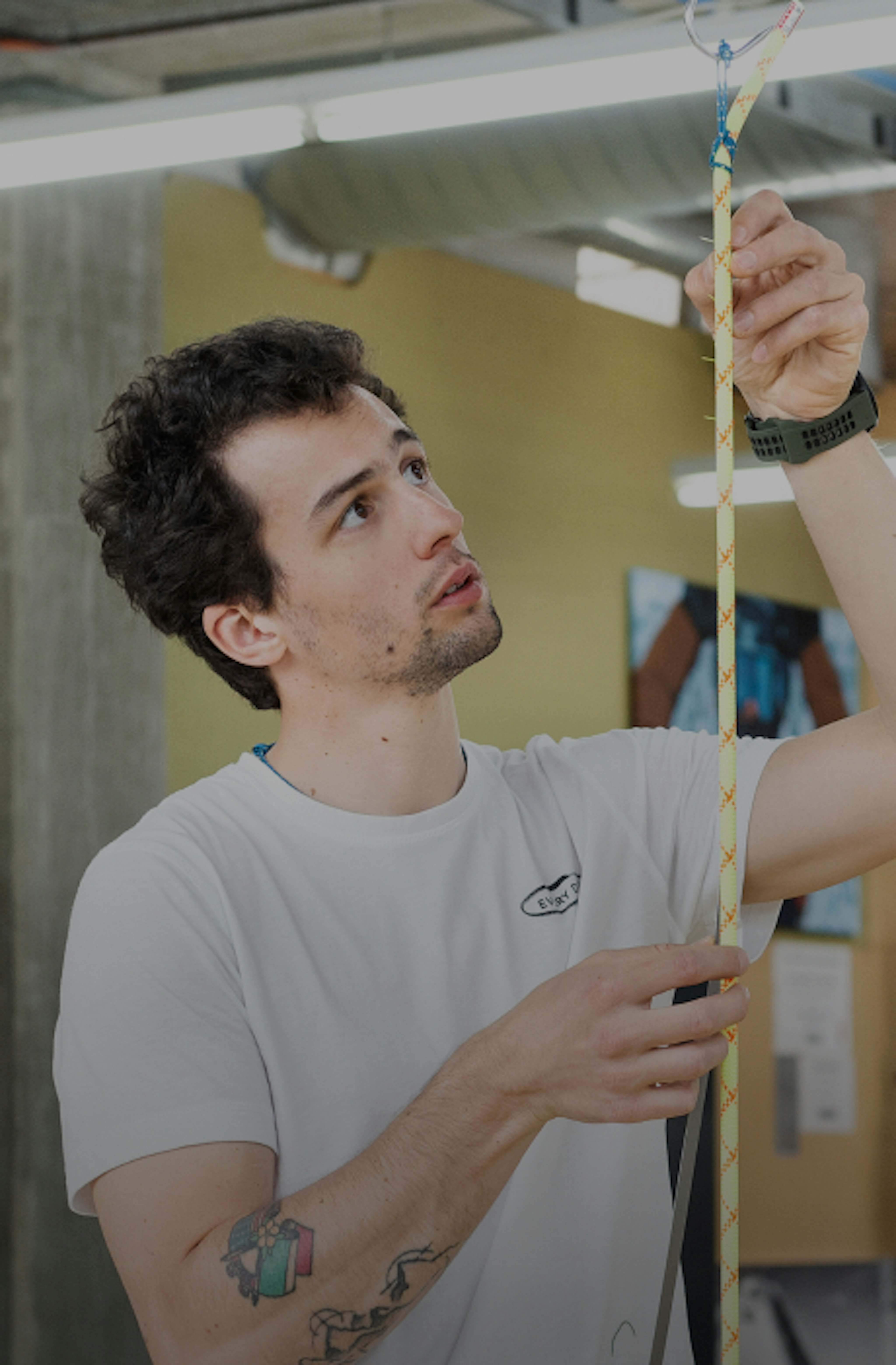 Person in a white Mammut shirt handling a patterned climbing rope indoors, looking up while adjusting the rope.