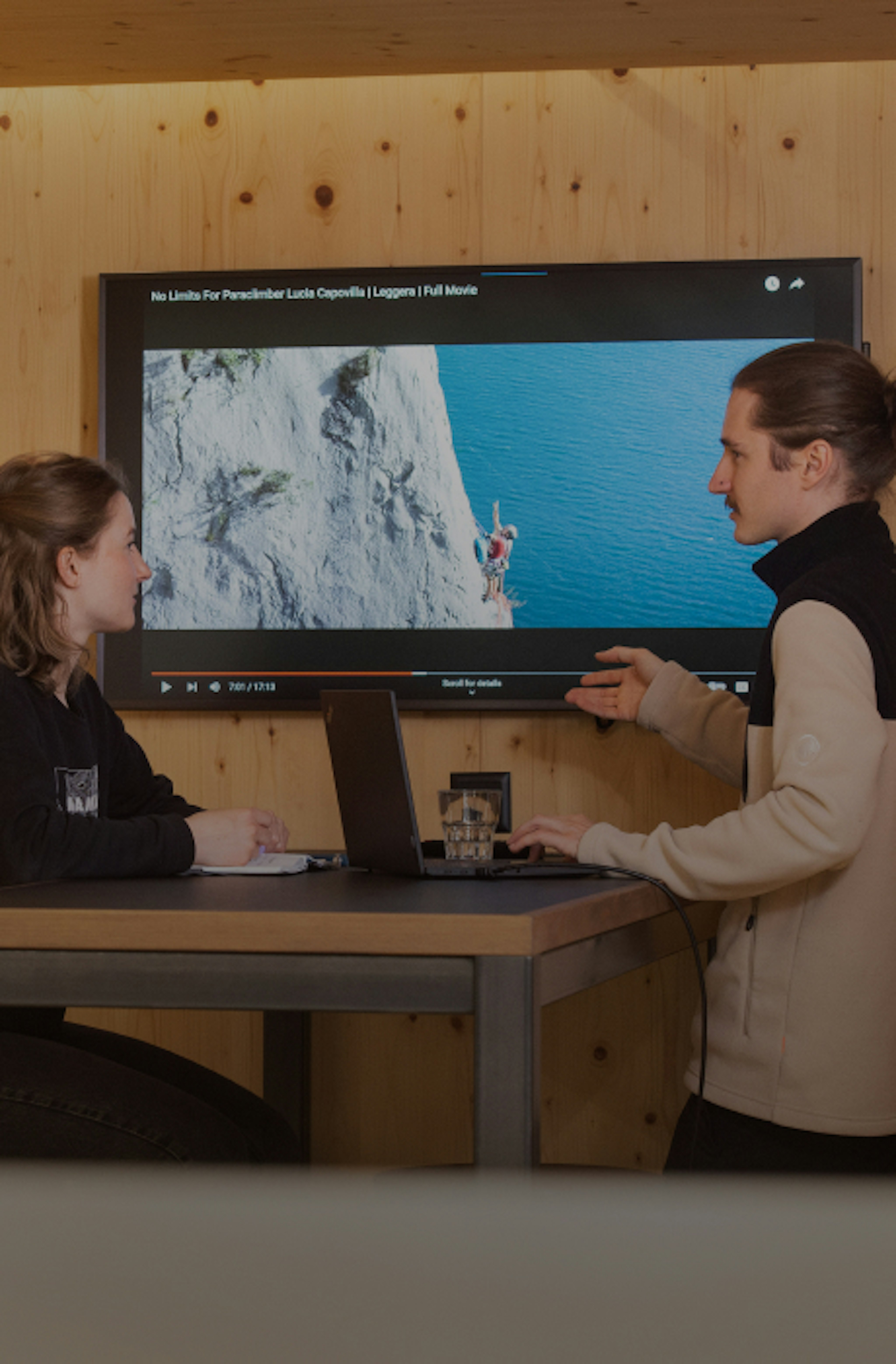 Two climbers sit at a table in Mammut gear, watching a climbing tutorial video on a screen mounted on a wooden wall.