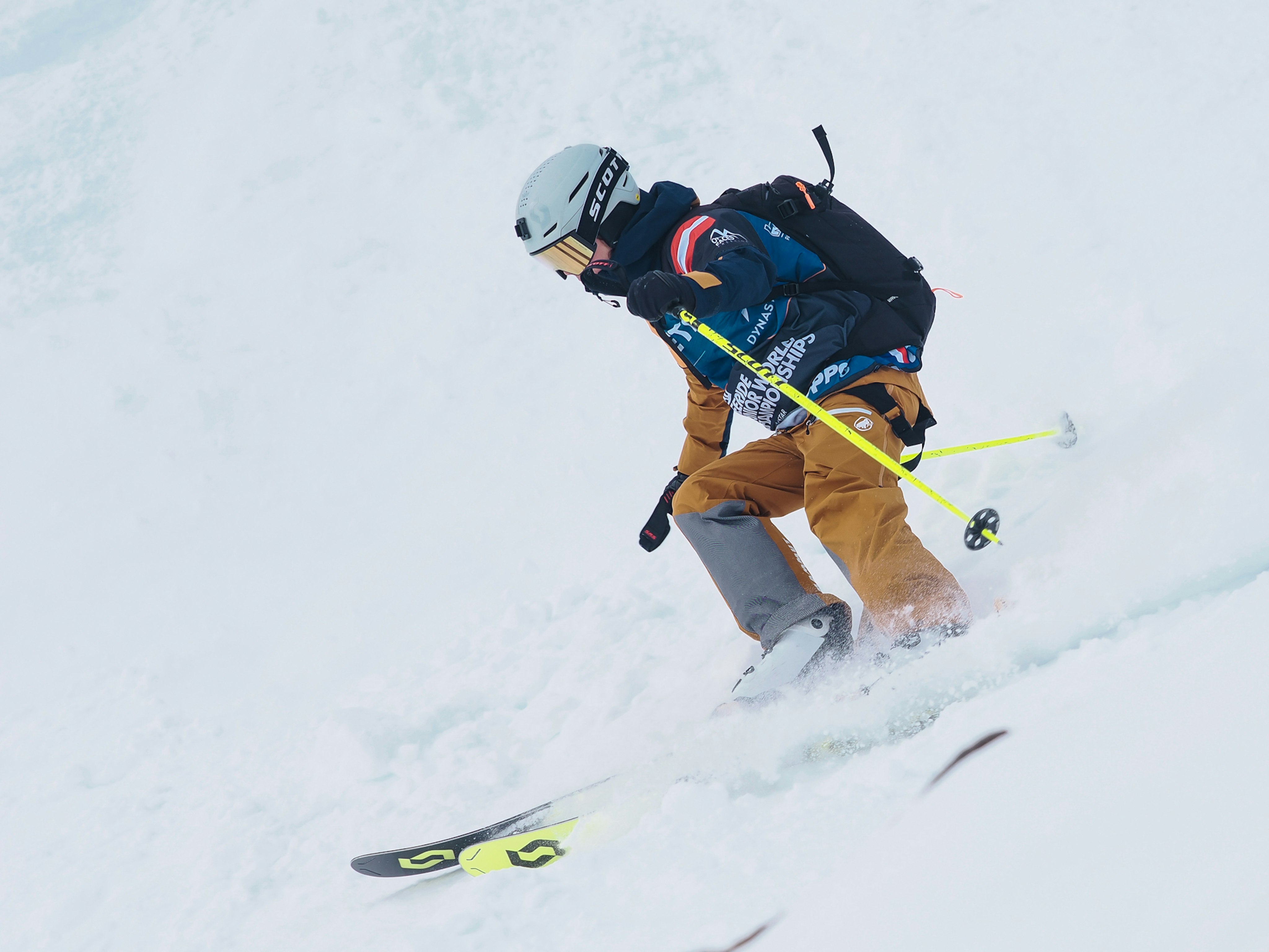 Zoe Delzoppo in Mammut gear navigating a snowy downhill slope, equipped with a helmet, goggles, ski poles, and a backpack.