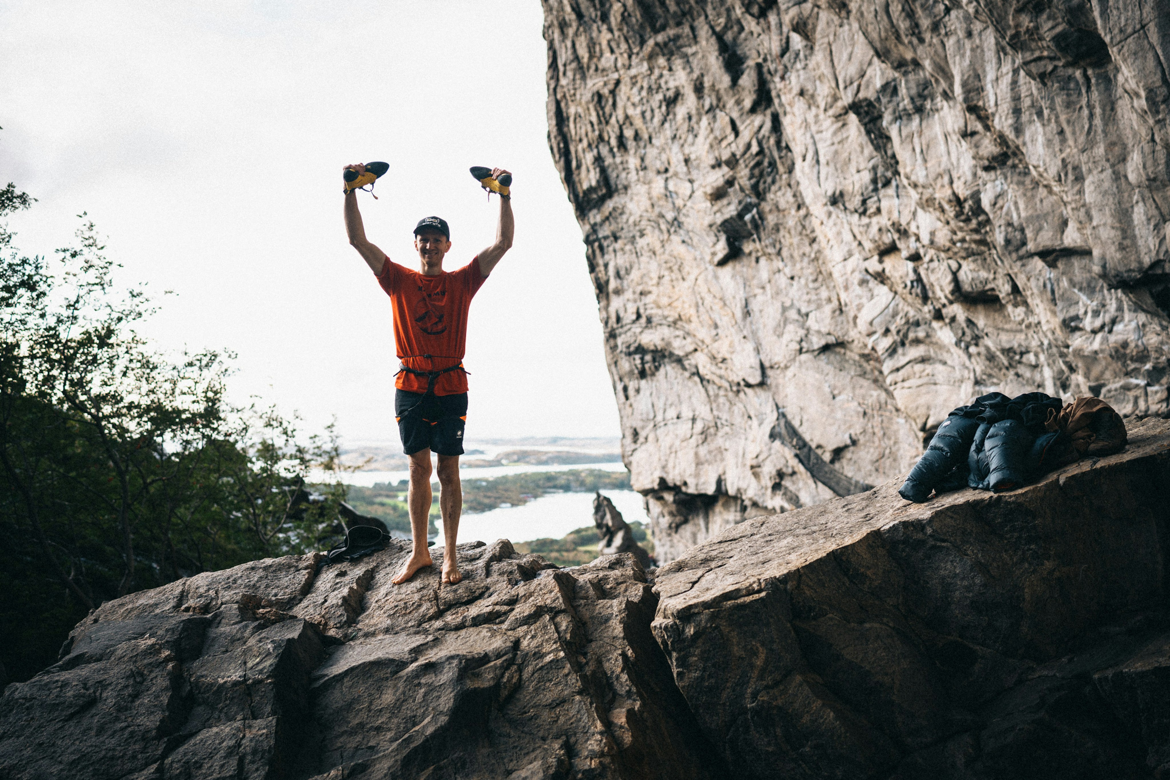 Jakob Schubert a climber in a Mammut orange shirt stands triumphantly on a rocky ledge, holding up Mammut climbing shoes, with a towering cliff and lush forest behind.