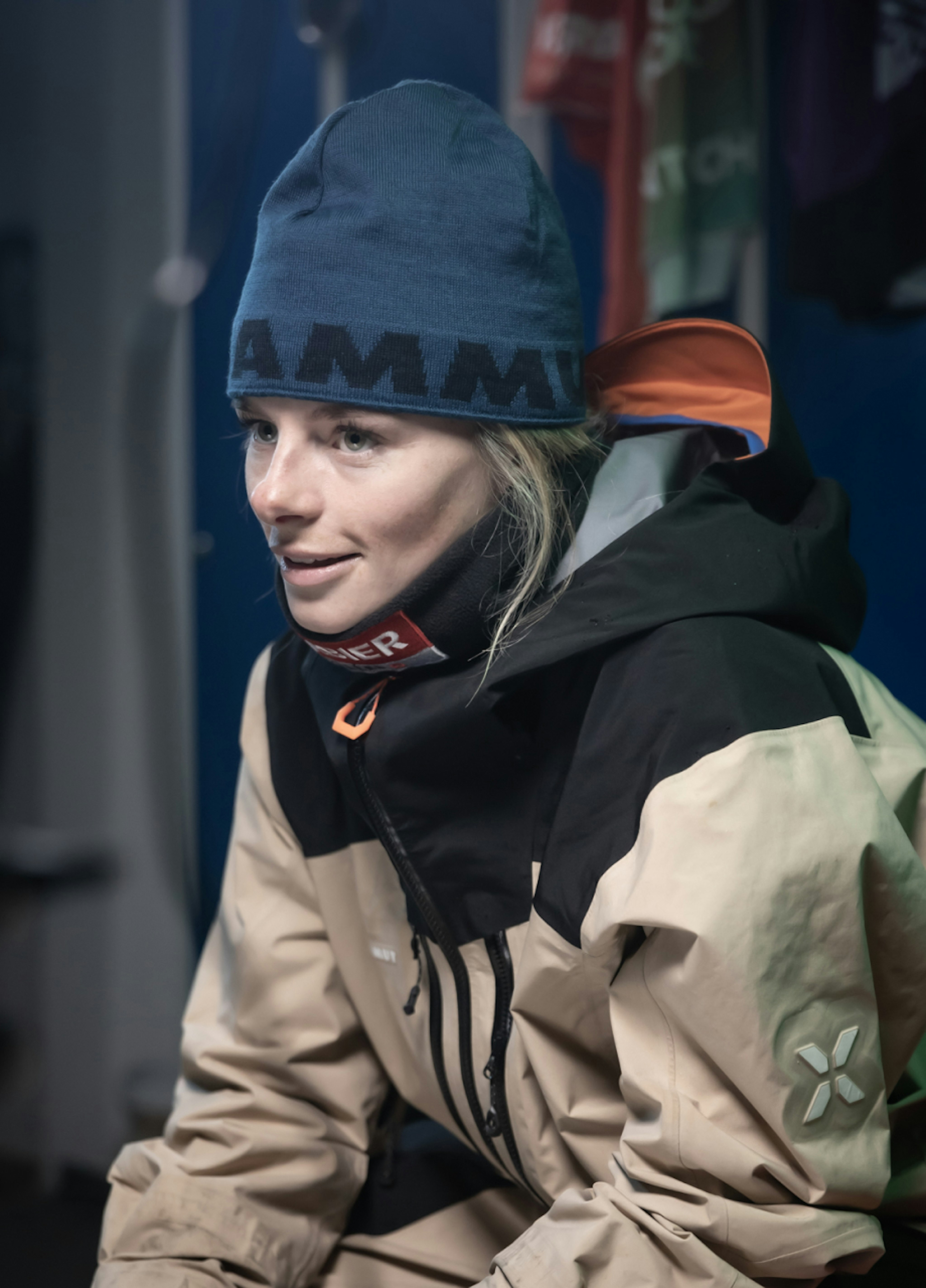 Person wearing a Mammut beanie and insulated jacket looking thoughtful, with sports jerseys blurred in the background.