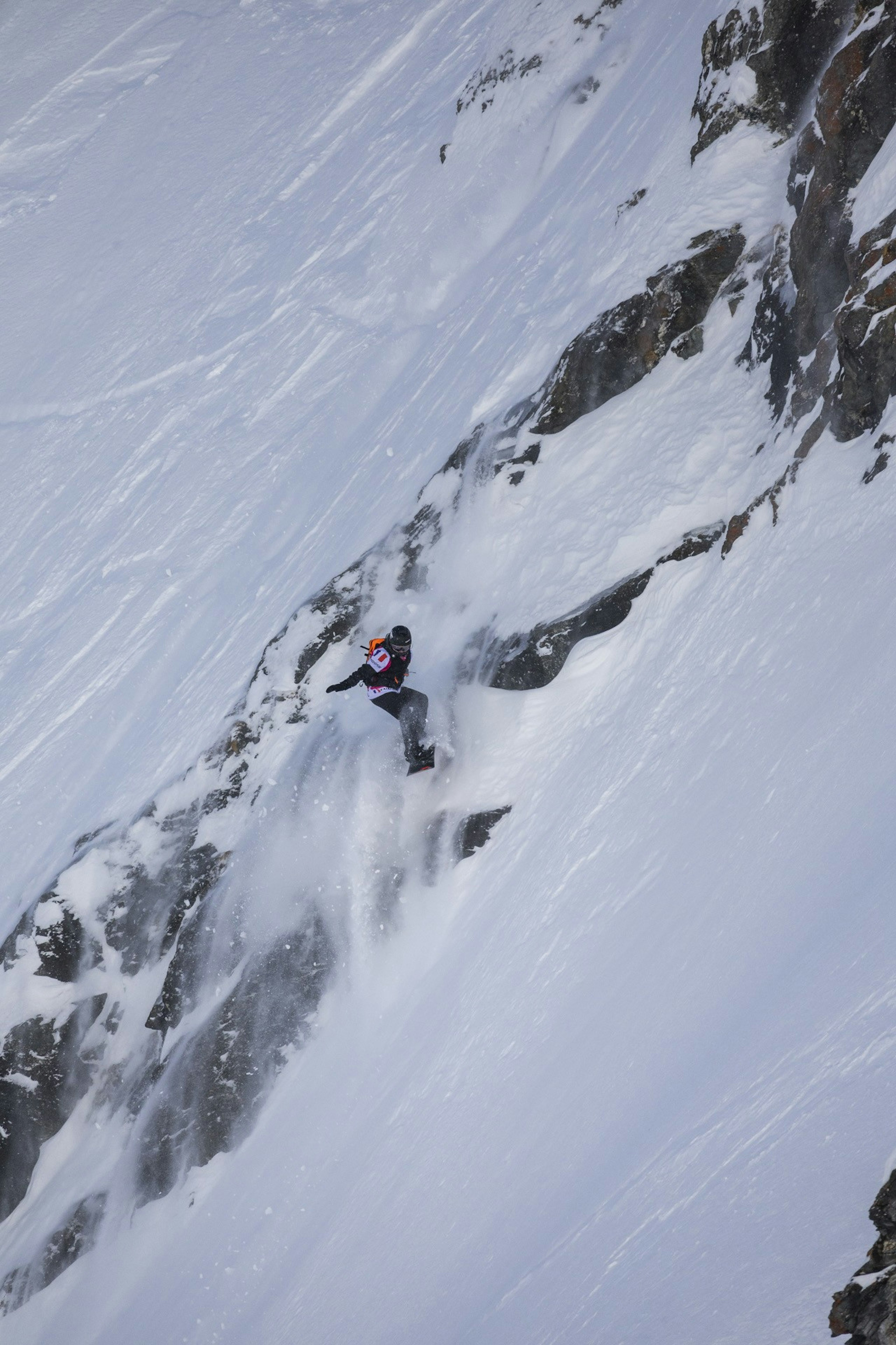 Mammut snowboarder expertly carving down a steep, rocky, snow-covered mountain slope with dynamic motion and impressive snow spray.