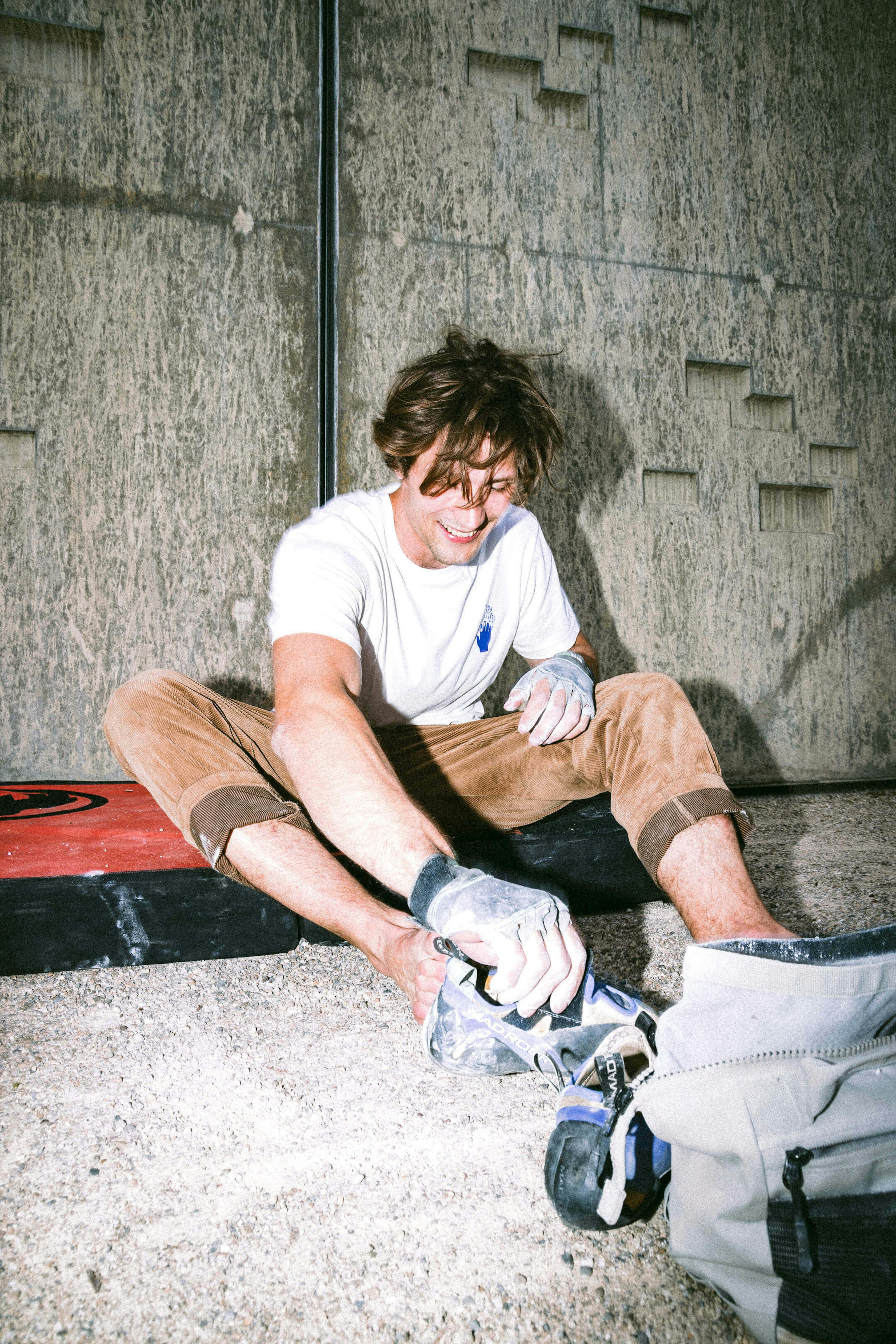 Person sitting on the ground, wearing Mammut climbing shoes and chalking hands, with Mammut climbing gear nearby and a climbing wall in the background.