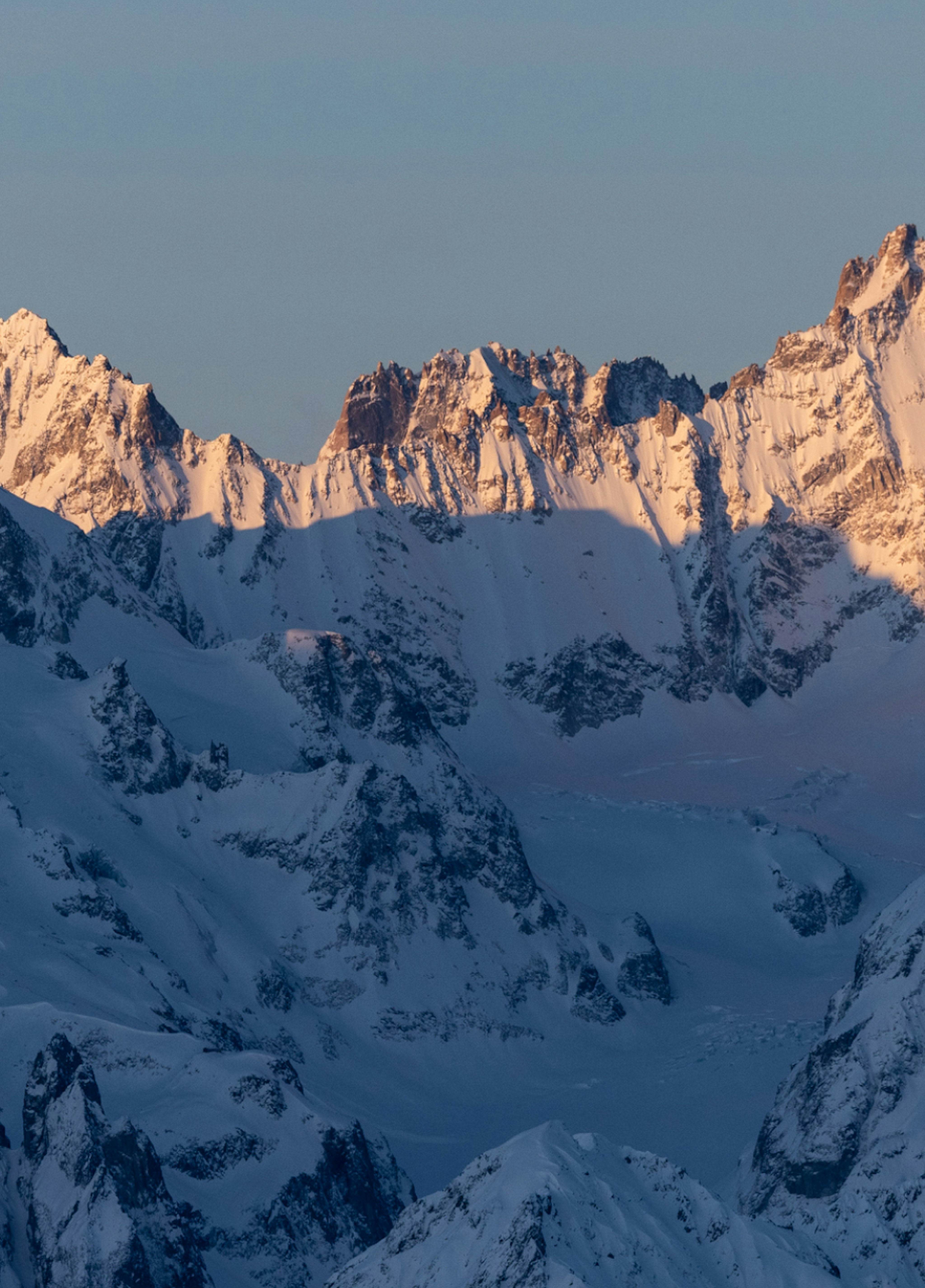 Snow-covered mountain range with peaks illuminated by the warm glow of the setting sun against a clear sky, perfect for Mammut mountaineering gear.