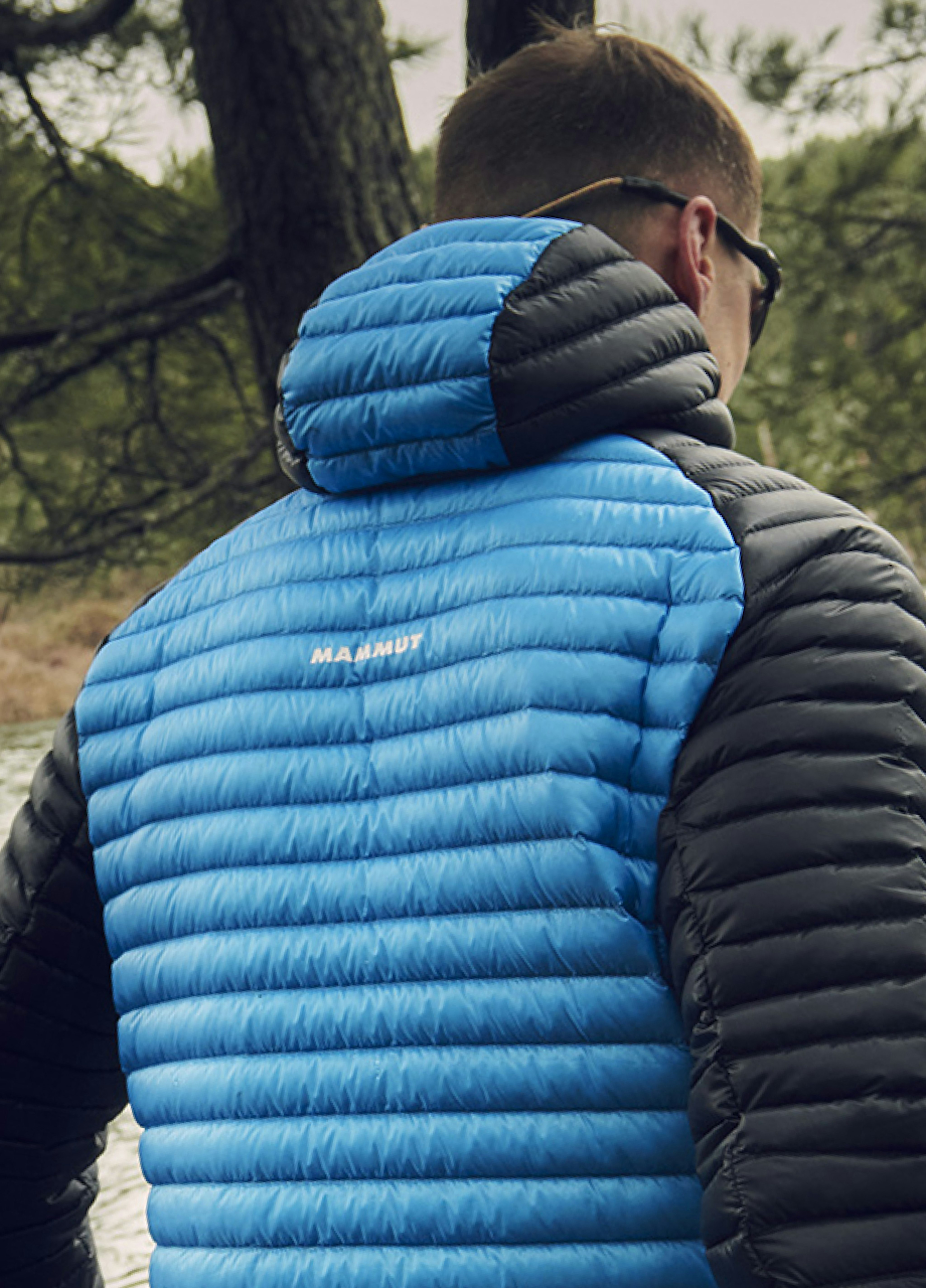 Man in a Mammut blue and black dry down jacket standing outdoors, near a tree, facing away from the camera.