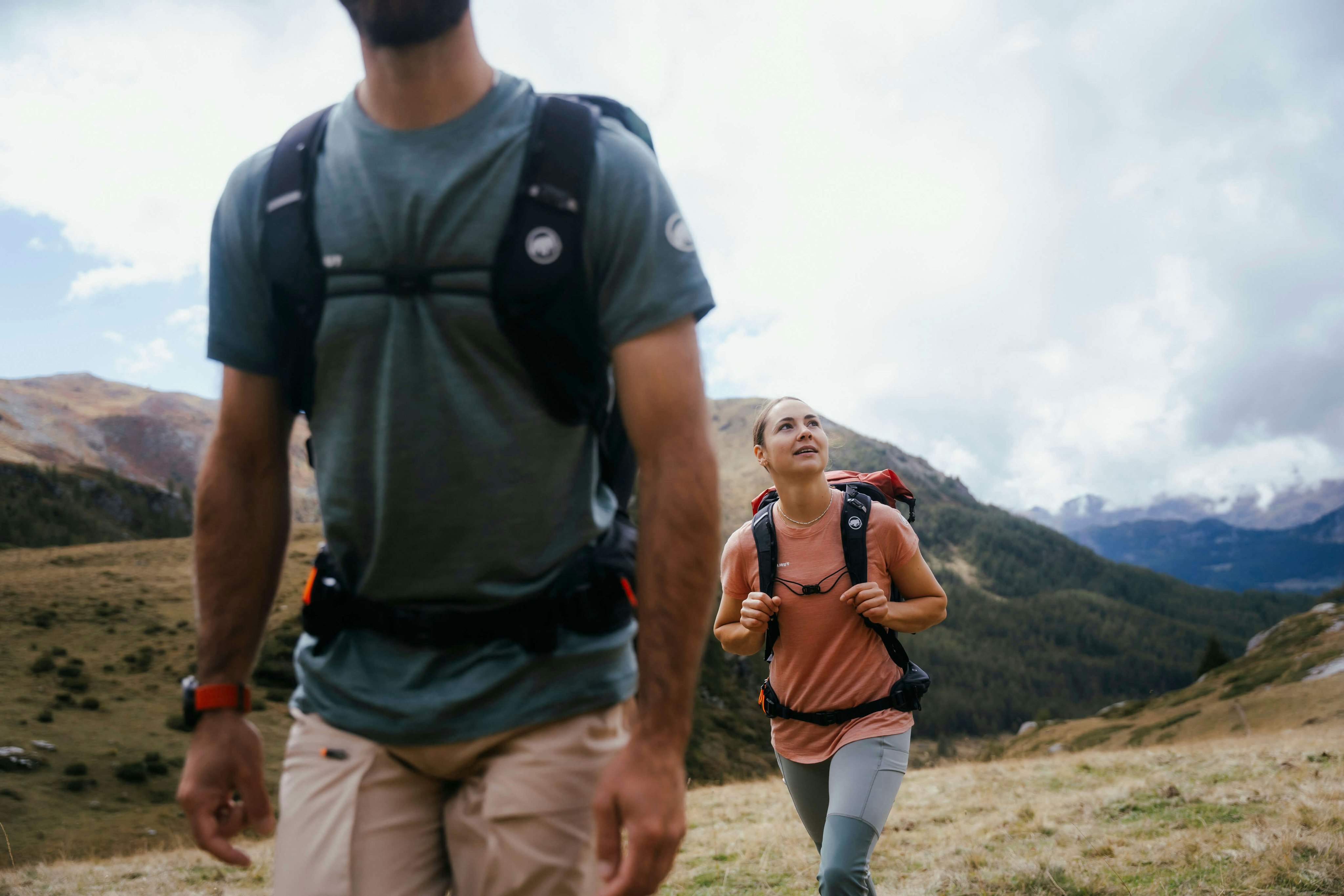 Two Mammut-equipped hikers with backpacks trek through a rugged mountainous landscape on a cloudy day, one in the foreground and one in the background.