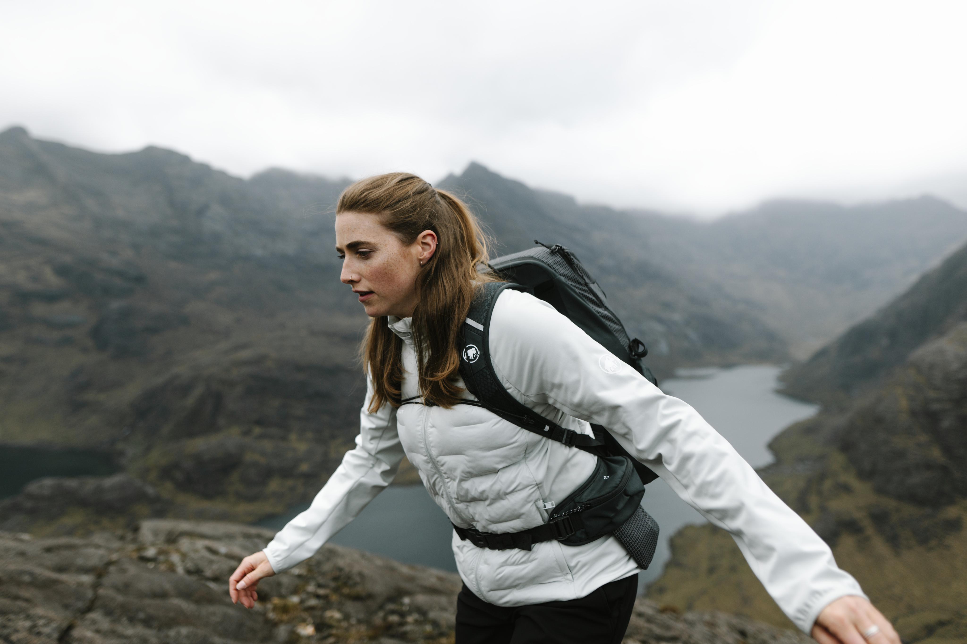 Smiling runner in Mammut sports gear and sunglasses rests outdoors, wearing a Mammut hydration vest and sun visor.