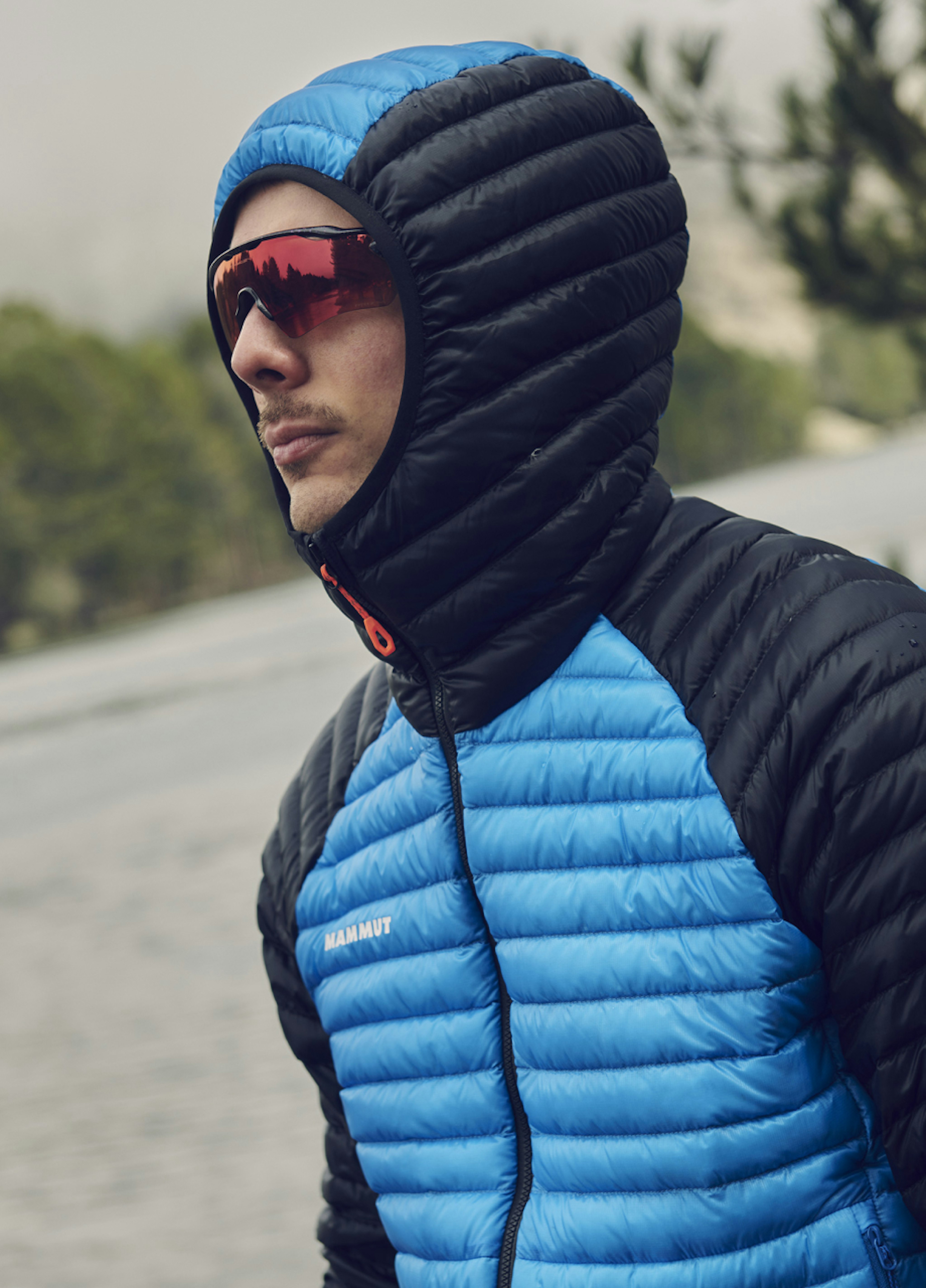 Person wearing a blue and black Mammut puffer jacket with a hood and red-tinted sunglasses, outdoors in a mountainous setting.