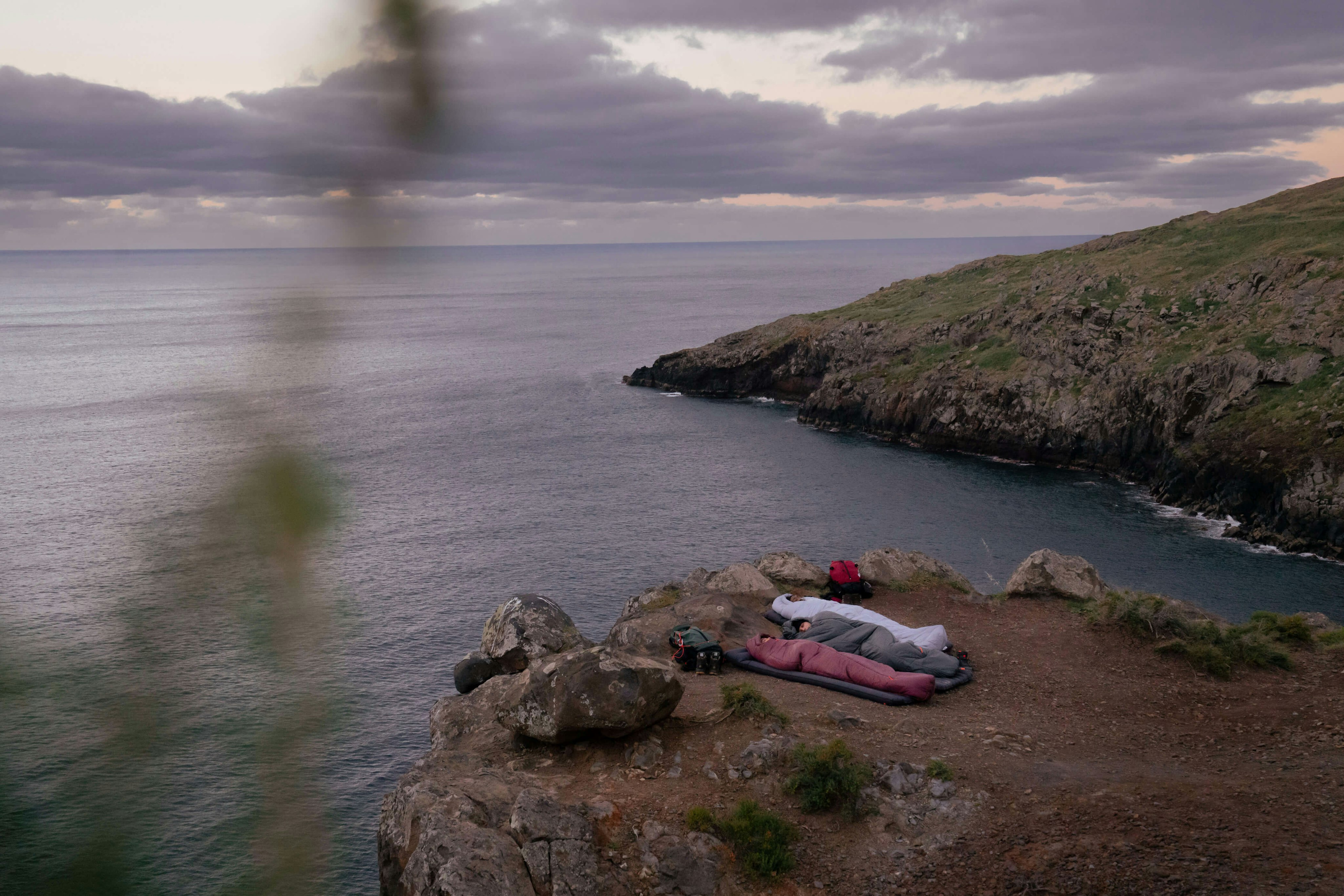 Person sleeping on a cliffside in a Mammut sleeping bag near the ocean, surrounded by rugged rocky terrain and an overcast sky.