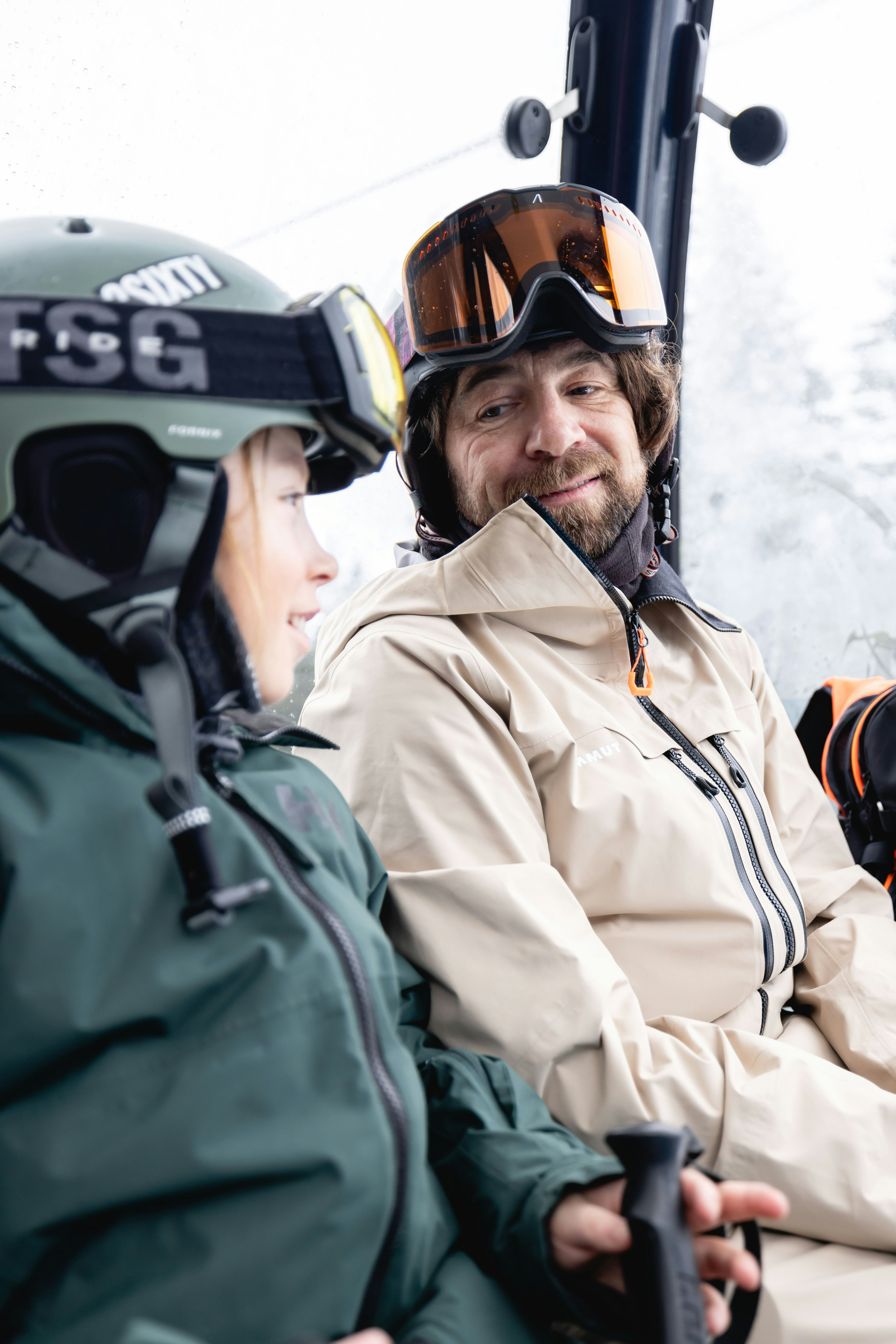 Man in Mammut ski gear with goggles smiles at a child inside a ski lift or gondola.