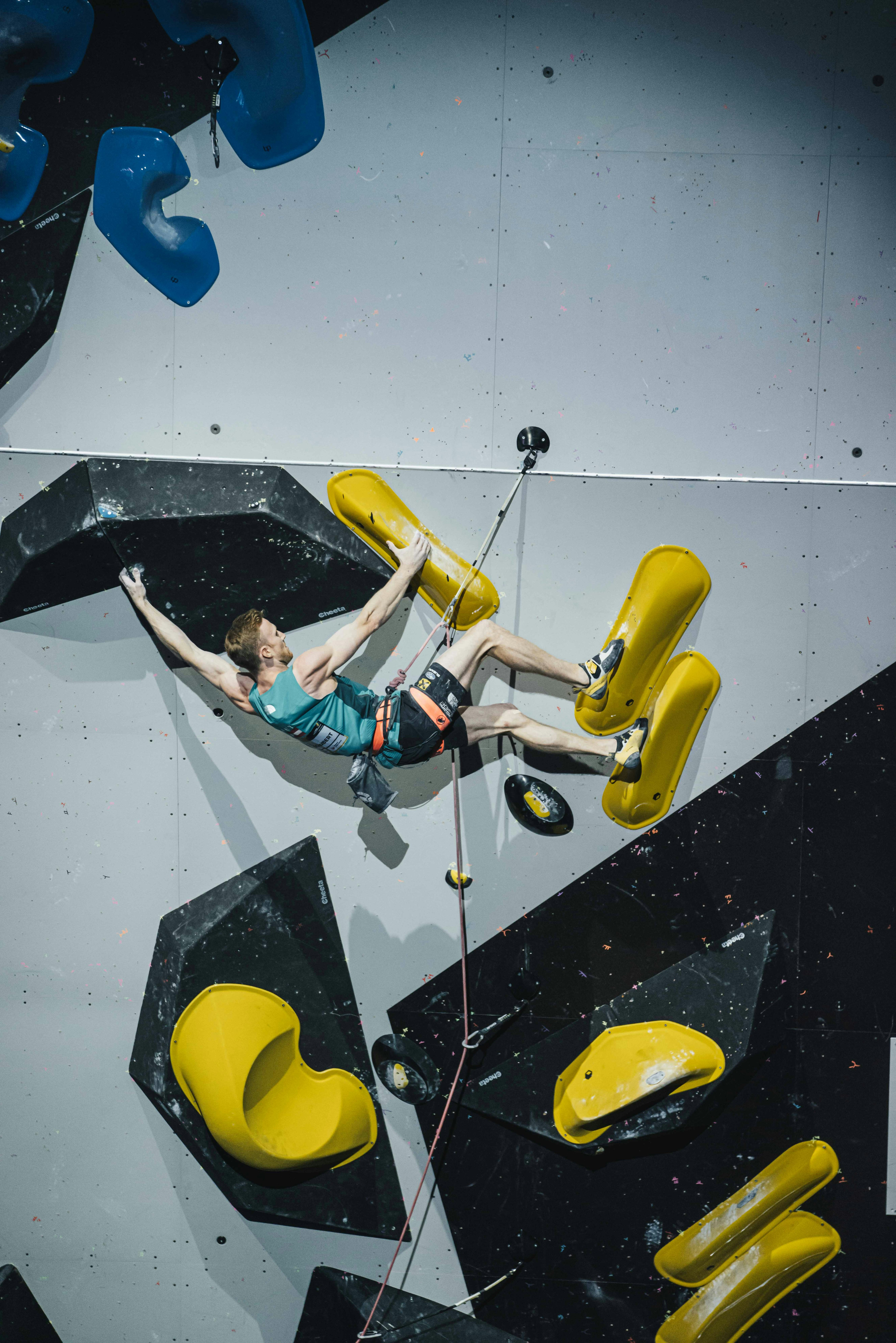 A climber in a blue Mammut tank top scales an indoor rock climbing wall with large yellow and black holds, showcasing their mountaineering equipment.