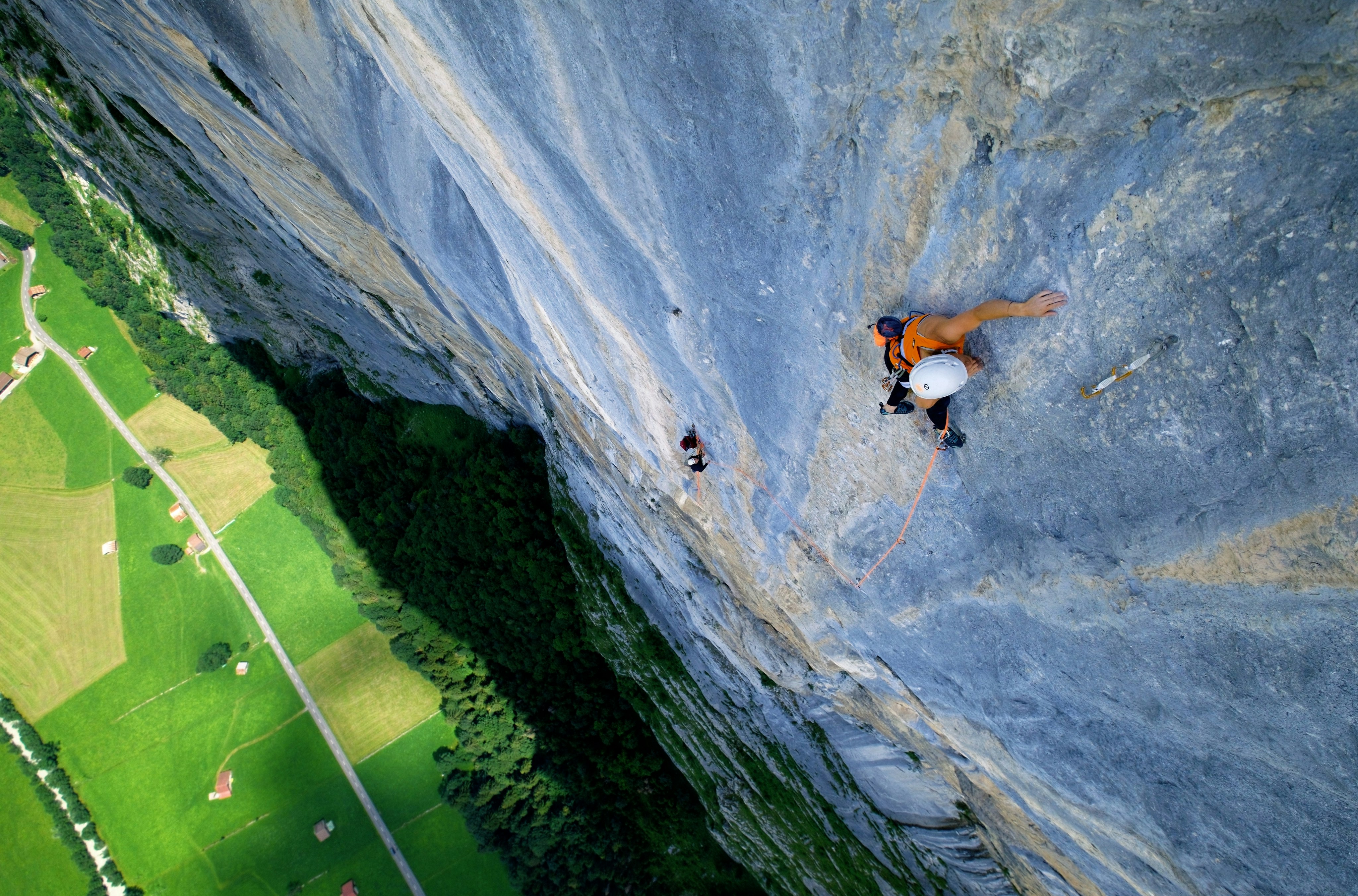 Katherine Chong, rock climbers in Mammut gear ascending a steep cliff face with a lush green valley below them. One climber is secured with a rope, showcasing outdoor adventure and safety.