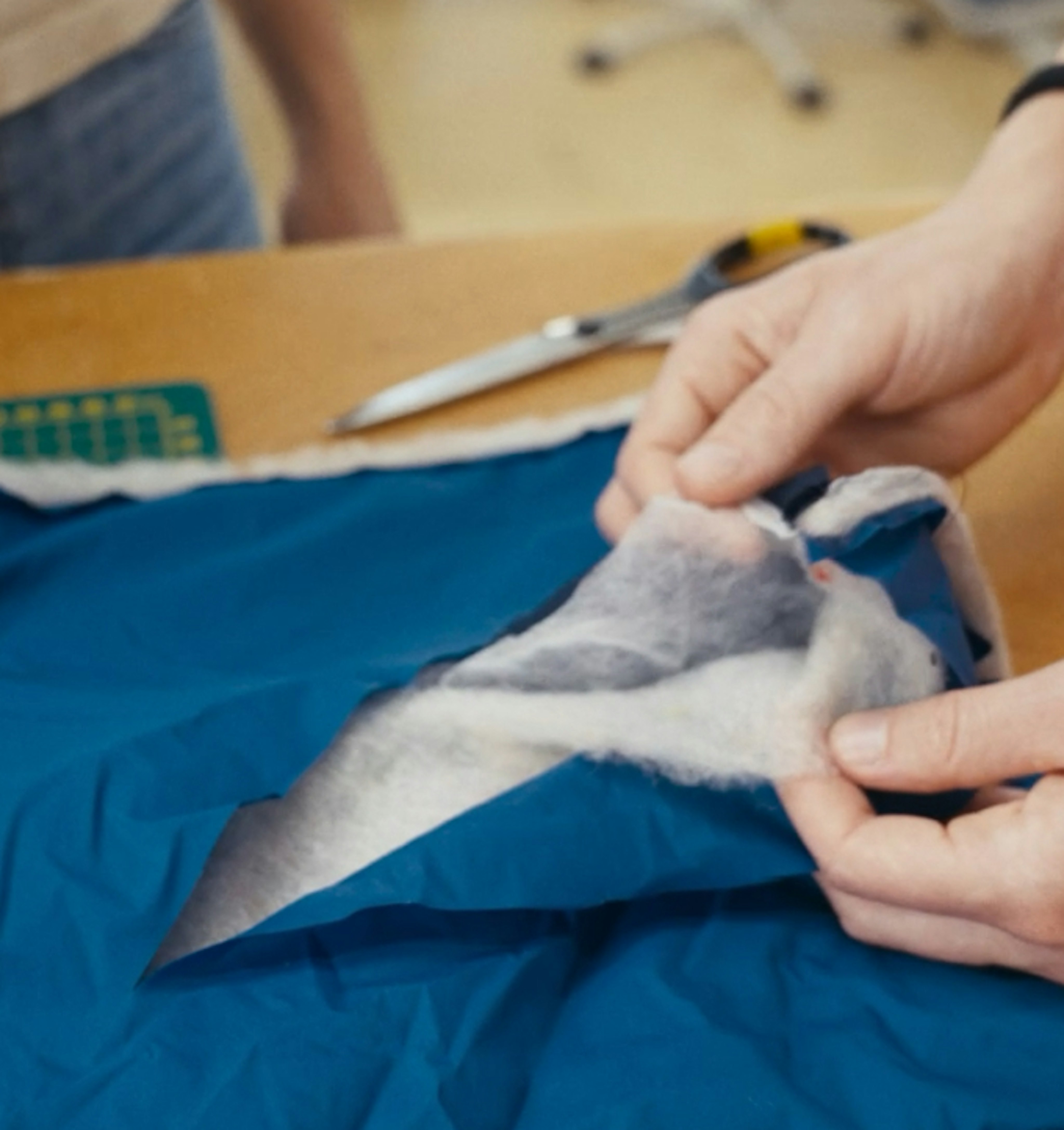 Hands crafting a blue Mammut mountaineering jacket, featuring fabric and stuffing, with scissors and a ruler in the background.