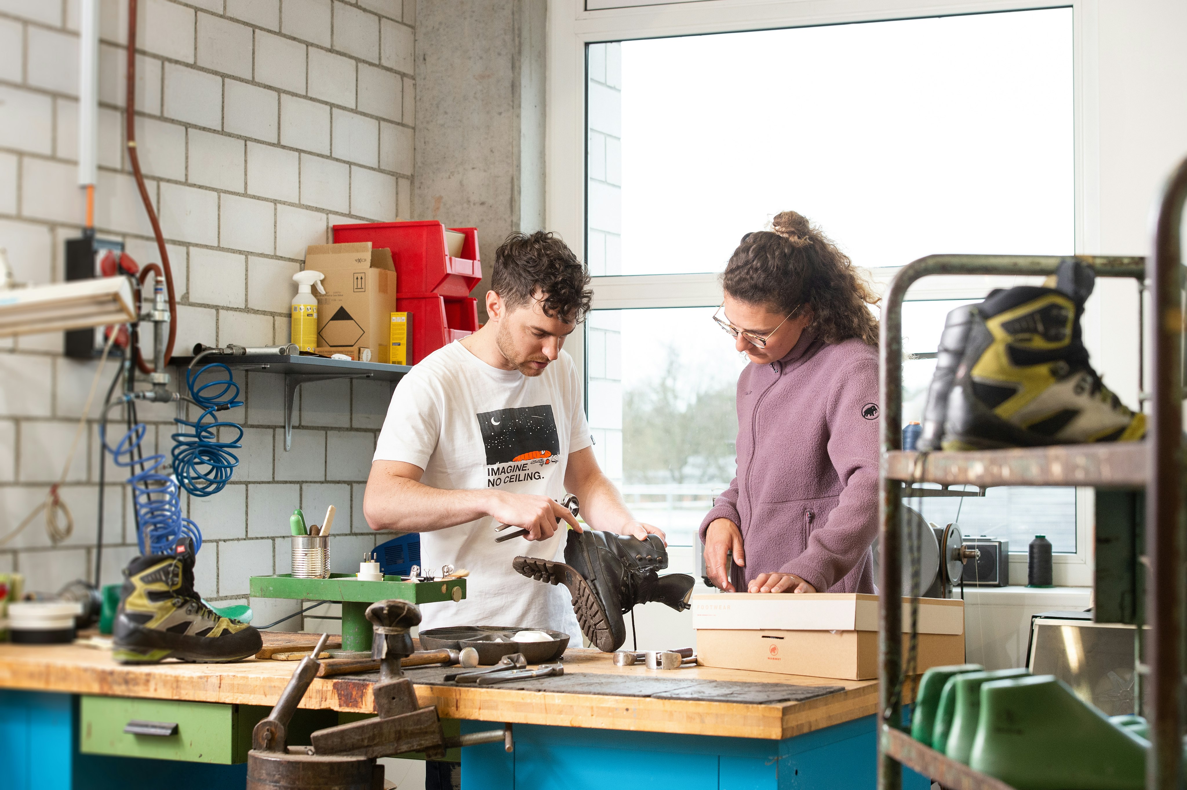 Two people repair Mammut hiking boots in a well-equipped workshop filled with tools and equipment, bathed in natural light from a nearby window.