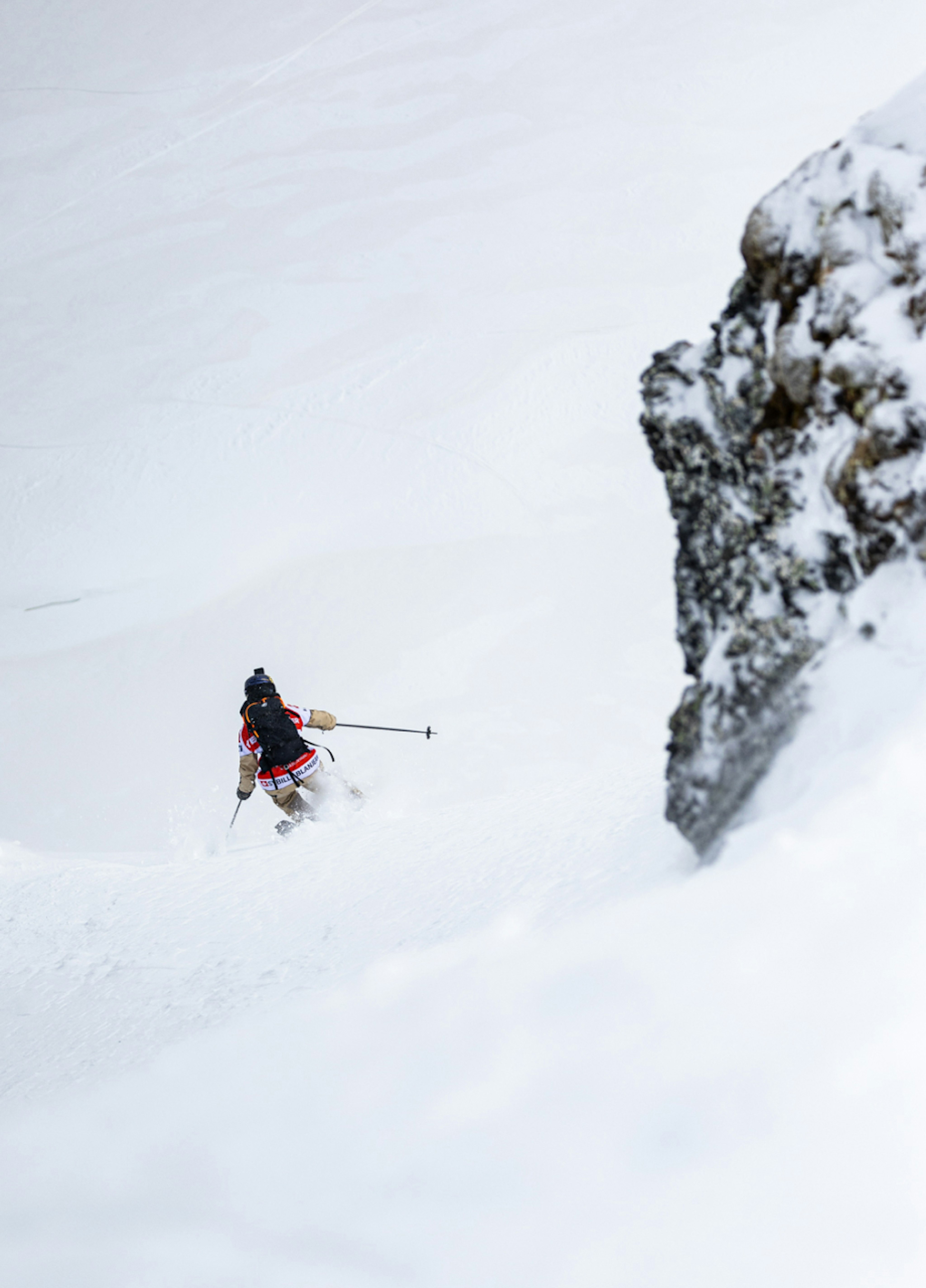 A skier in mid-descent carves through fresh snow on a steep slope, wearing Mammut gear, with a rugged rock face on the right and a pristine snowy landscape unfolding ahead.