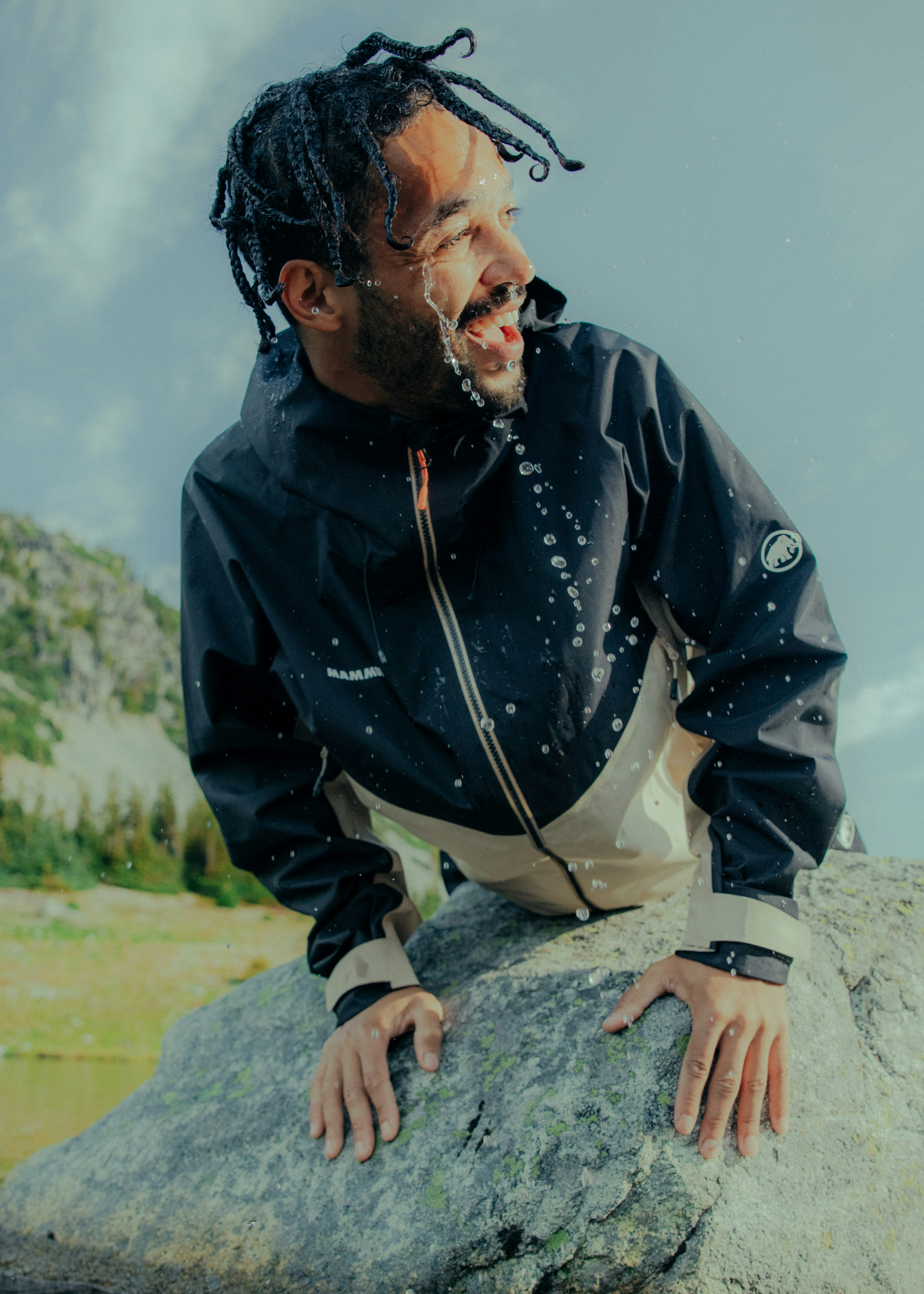 Person in a black and beige Mammut jacket smiles while leaning on a rock with water splashing in a natural outdoor setting.