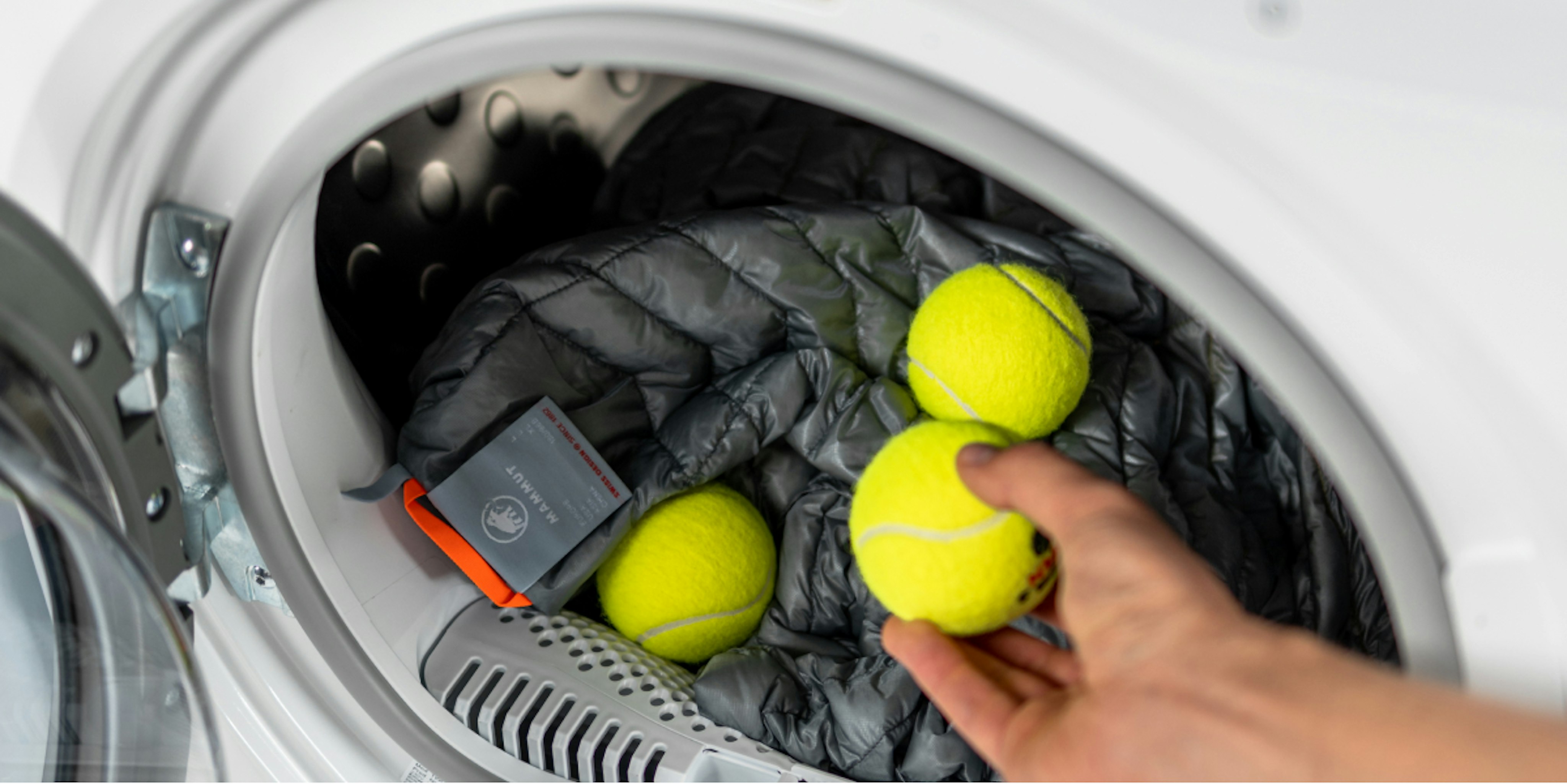 Hand placing tennis balls on a gray padded Mammut sports jacket inside an open washing machine.