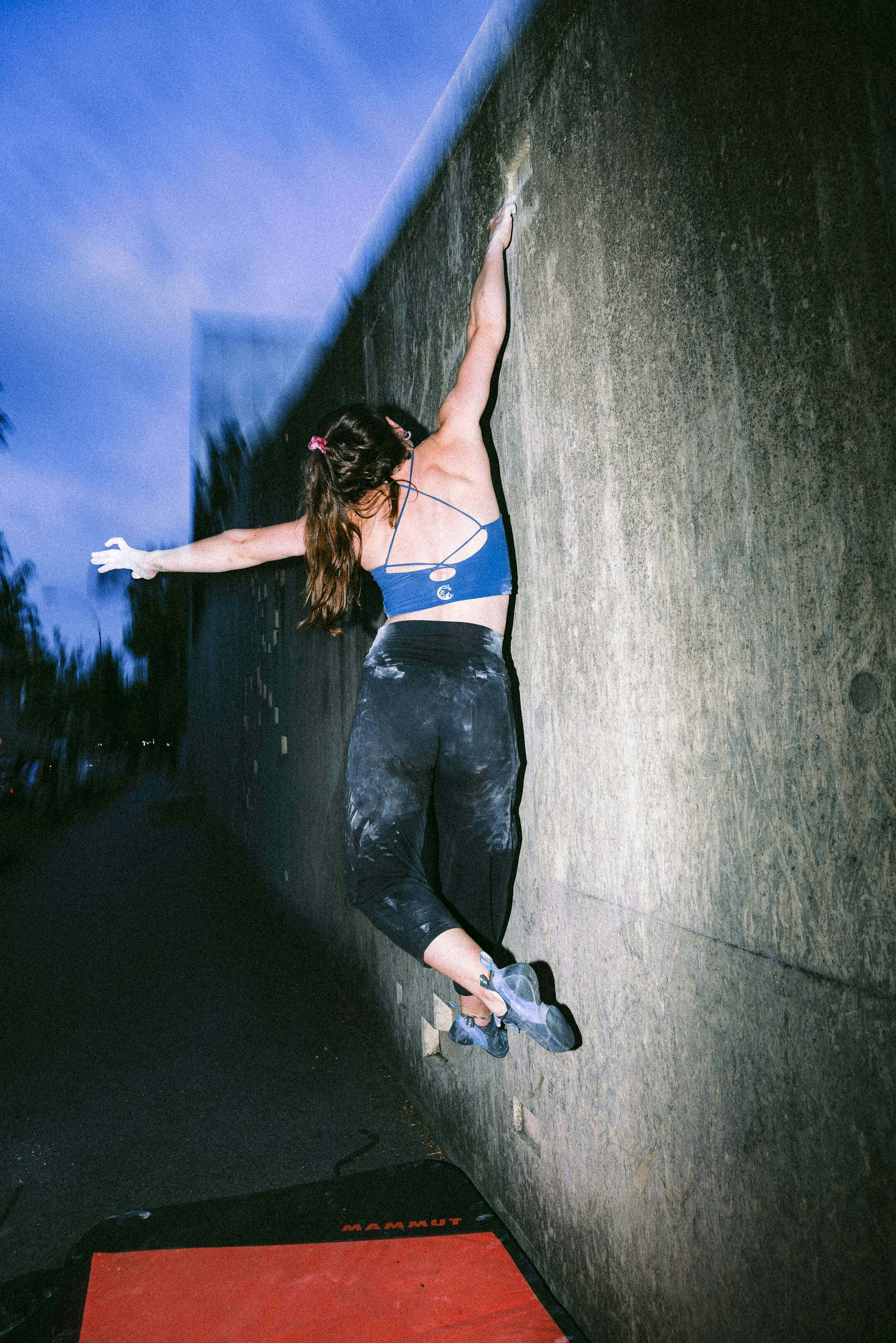 A climber in Mammut athletic wear scaling an outdoor climbing wall, with a crash pad below, during the early evening.