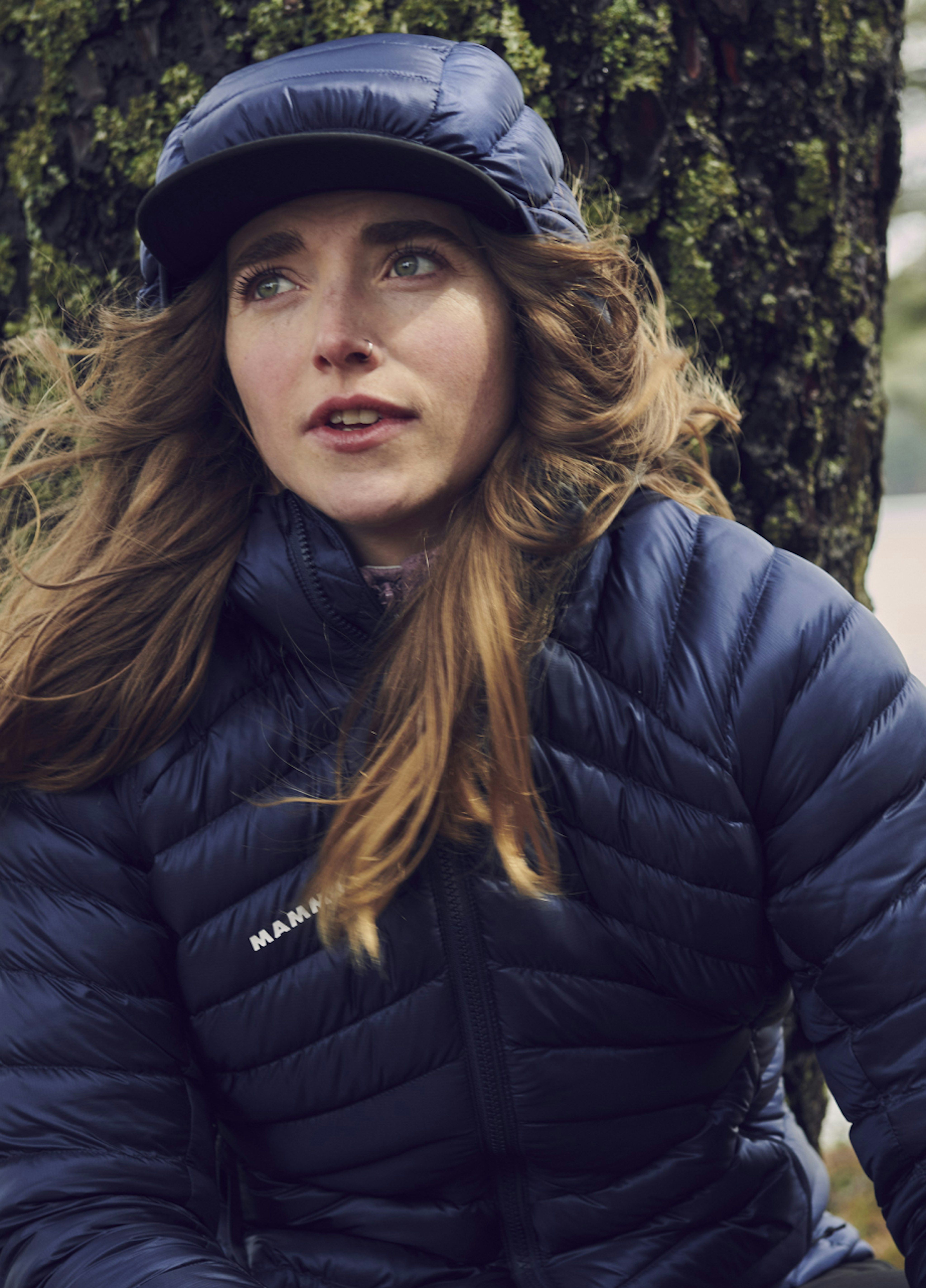 Woman in a Mammut navy dry downjacket and cap, leaning against a tree with windblown hair, looking thoughtful during an outdoor adventure.