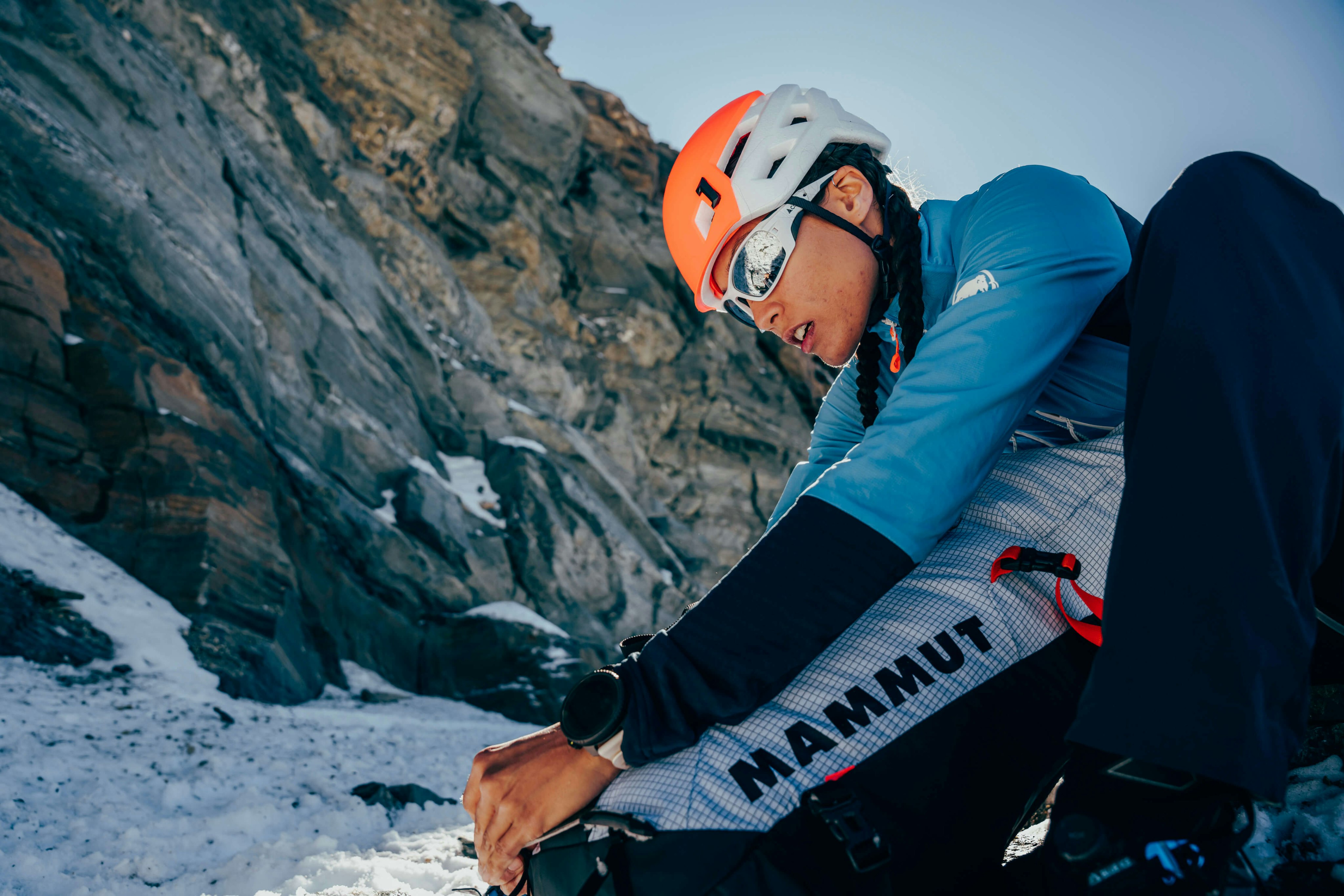 A climber wearing a Mammut helmet and glasses adjusts climbing gear on a snowy mountain, showcasing high-quality Mammut mountaineering equipment.